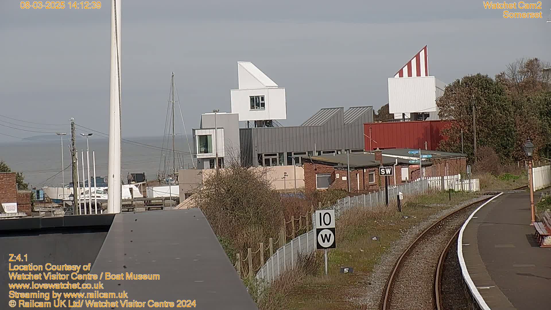 A view of a harbor with boats and modern buildings, including a distinctive white and red striped structure, on a sunny day, overlooks a railway track.