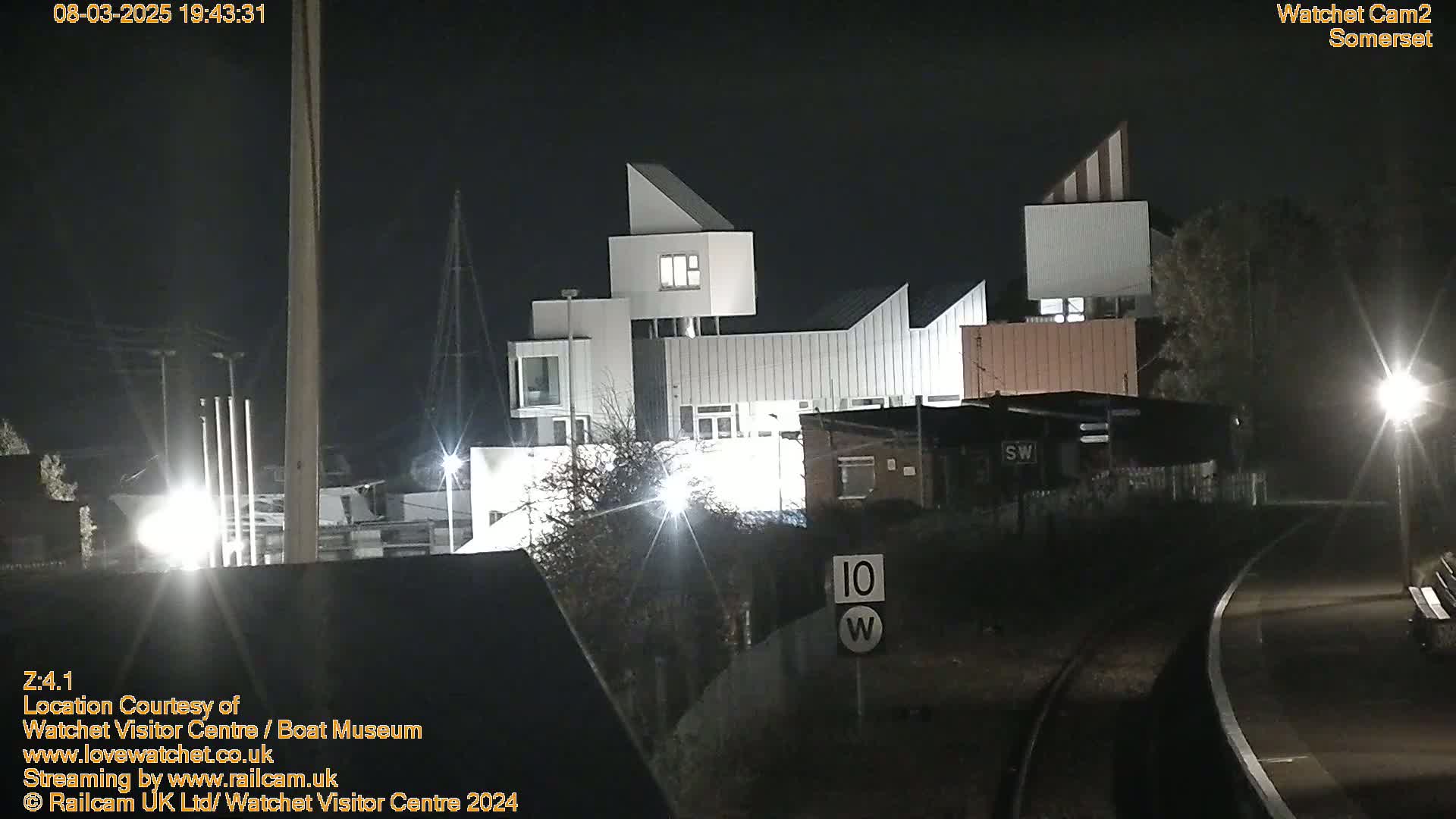 A modern, multi-level white building is illuminated at night, next to railroad tracks and other smaller structures.