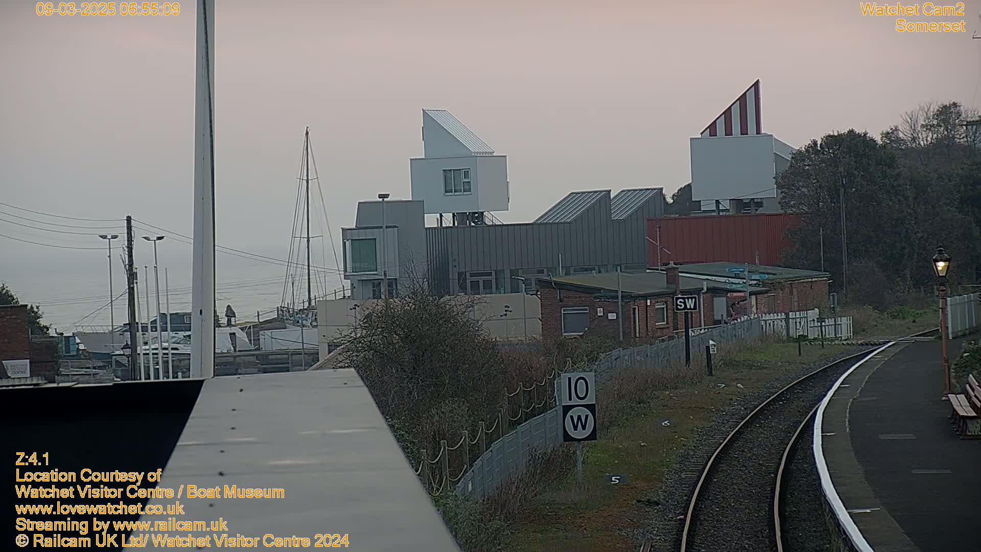A view of a harbor with modern buildings, a sailboat, and railway tracks under an overcast sky.