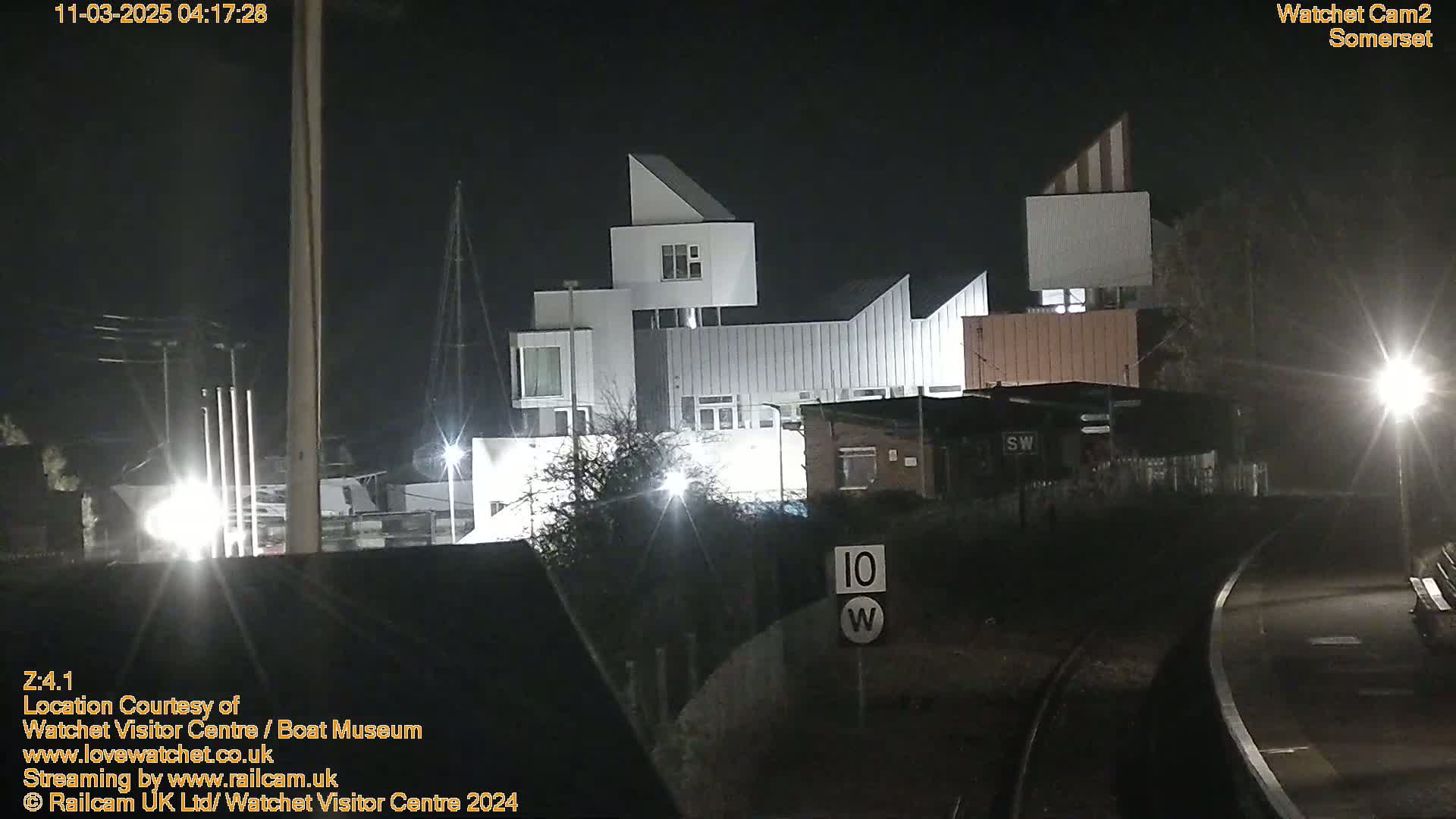 A nighttime view of a modern building complex near a roadway and railroad tracks under clear skies.