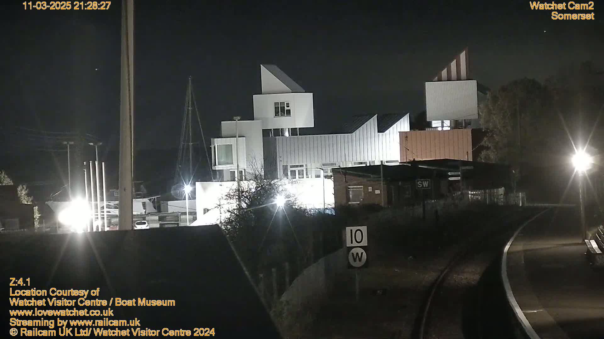 At night, brightly lit modern buildings are situated near train tracks under a clear, dark sky.
