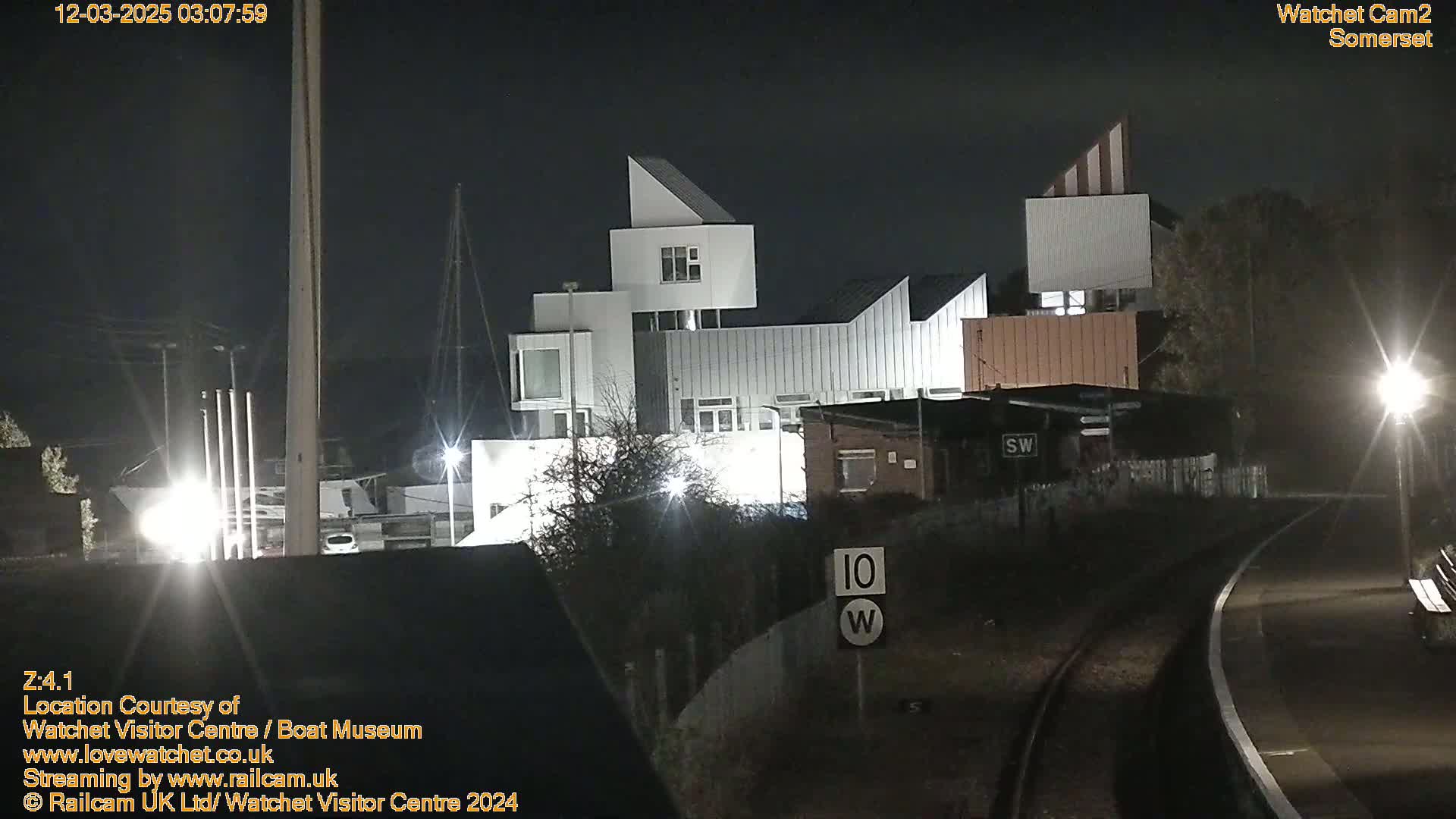 At night, a modern building complex is illuminated, situated near a roadway and railway tracks.