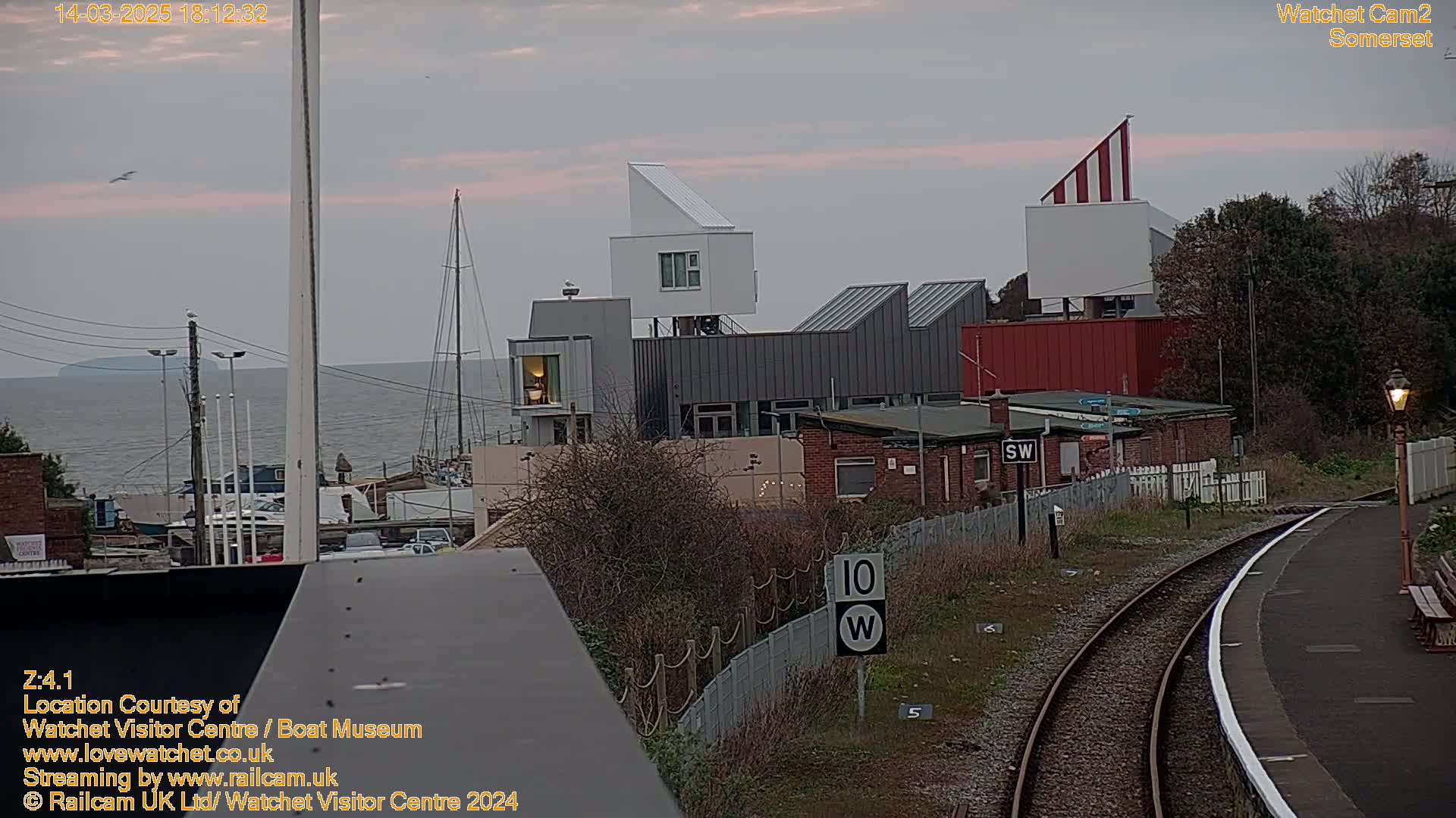 A modern, multi-level building complex sits near a train station and a calm sea under an overcast sky.