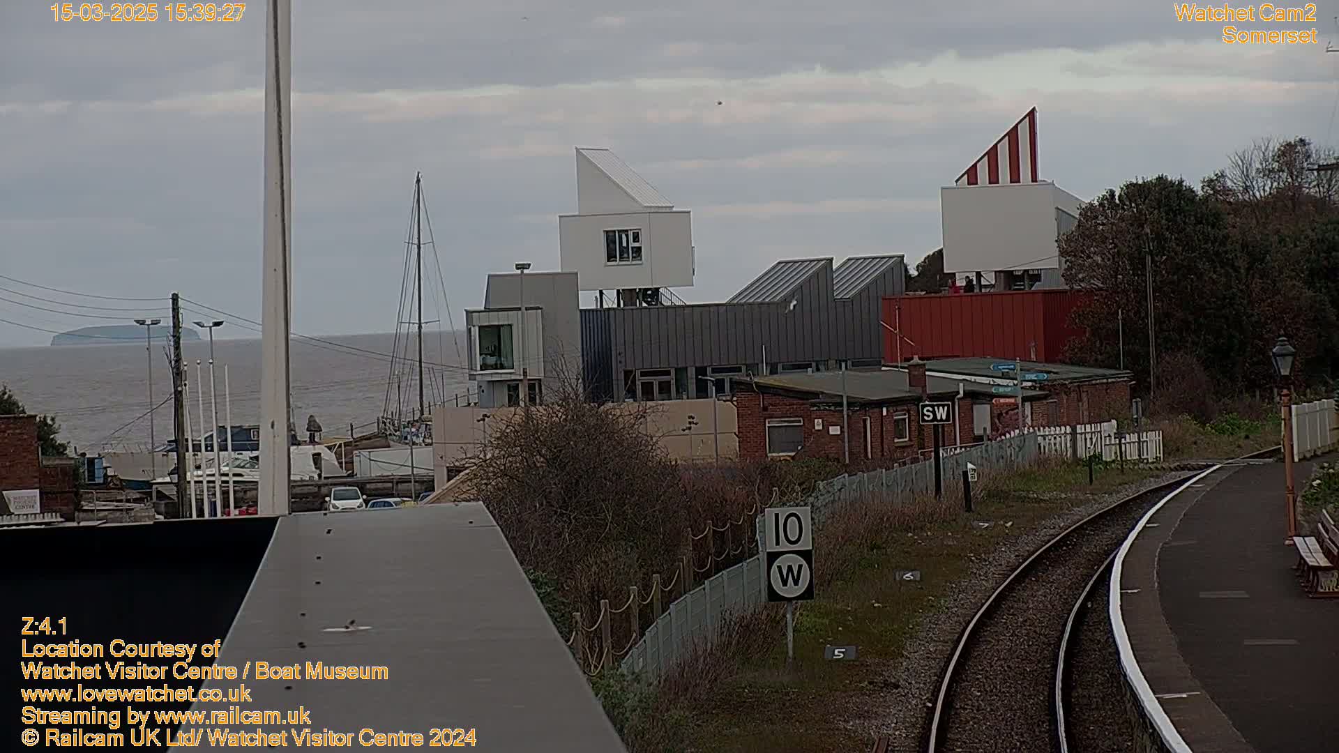 A view of a waterfront area with modern buildings, a sailboat, and train tracks under an overcast sky.