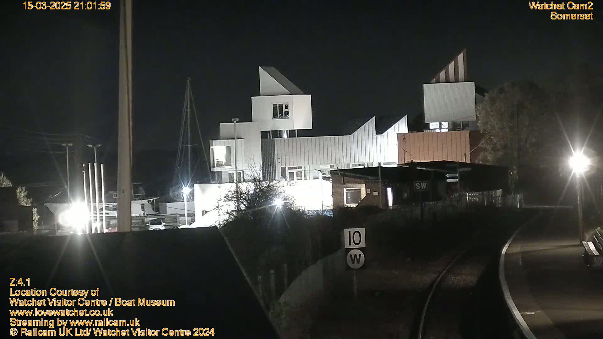 A modern, angular building complex is illuminated at night near a railway line under a dark sky.