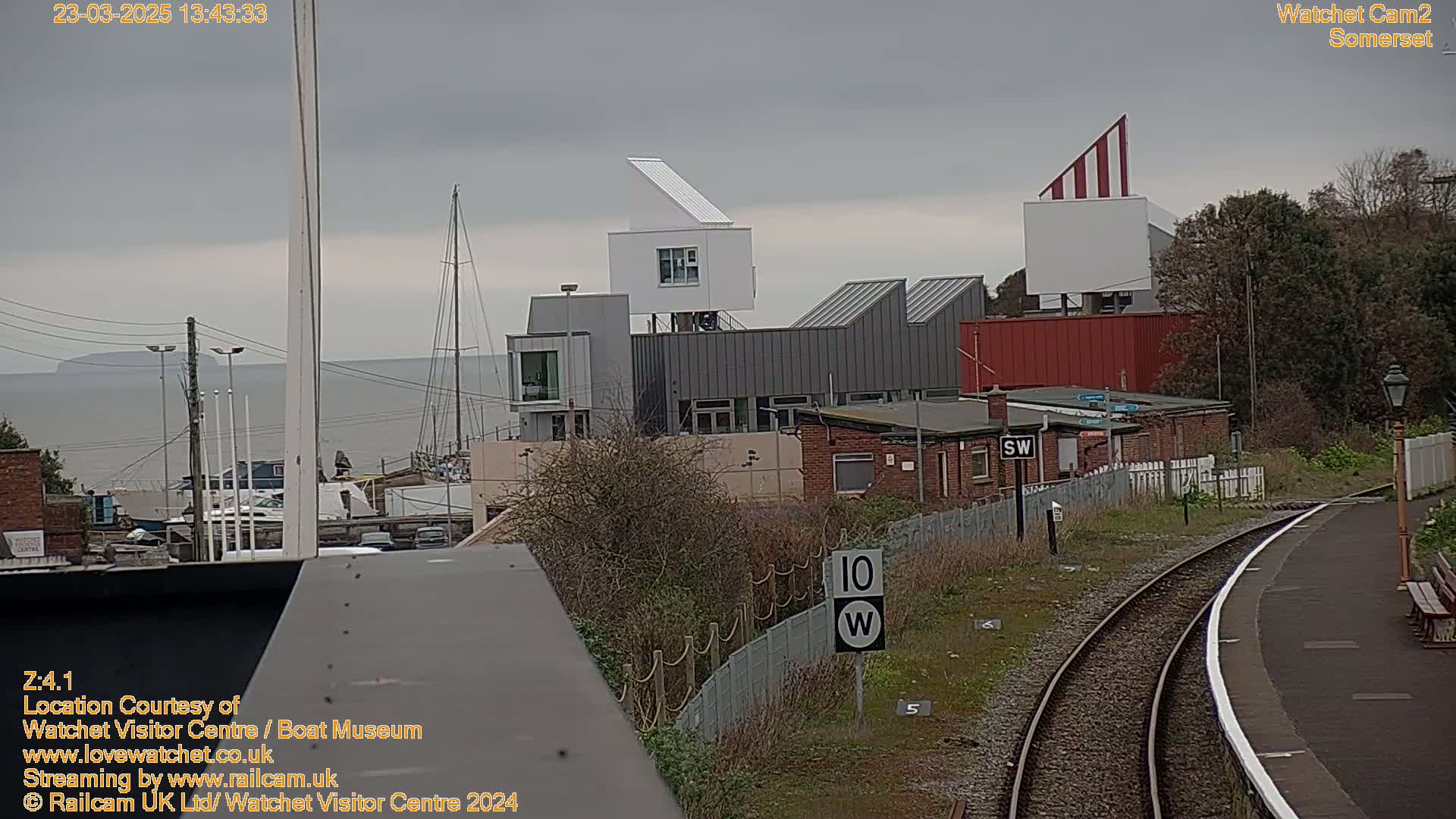 A view of a harbor with modern buildings, a sailboat, and train tracks under an overcast sky.