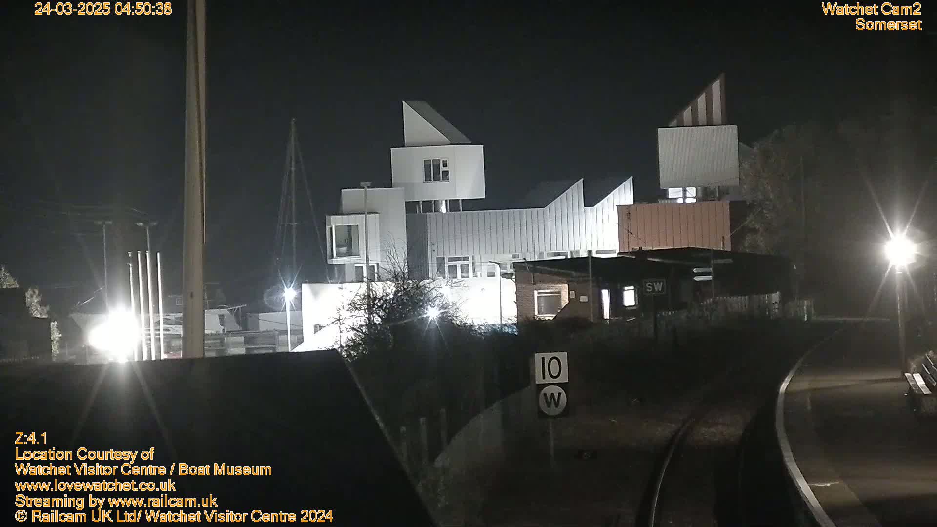 At night, a modern white building complex is illuminated near train tracks under a dark sky.