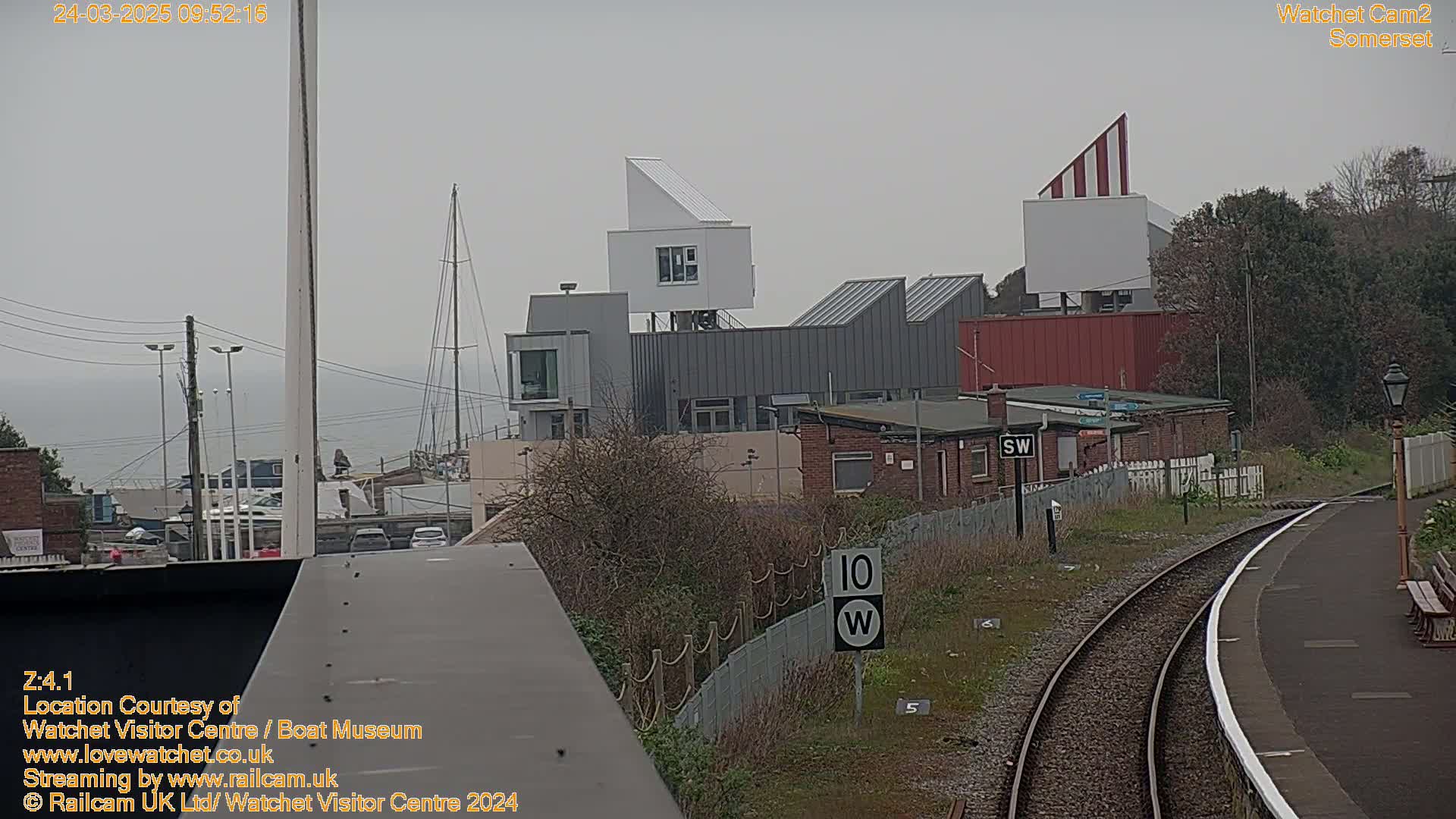 A view of a harbor with modern buildings, a sailboat, and a railway track on an overcast day.