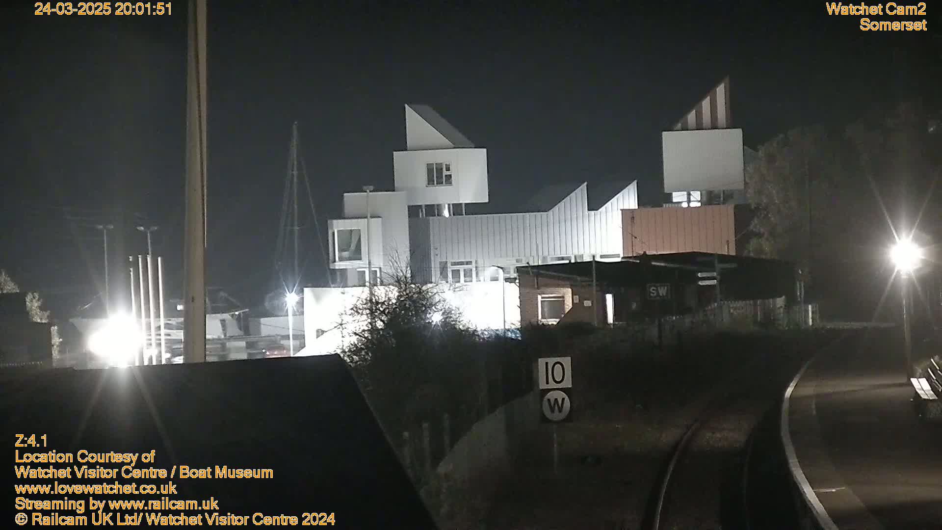 A modern, multi-story building is illuminated at night, near a railway track with a speed limit sign.