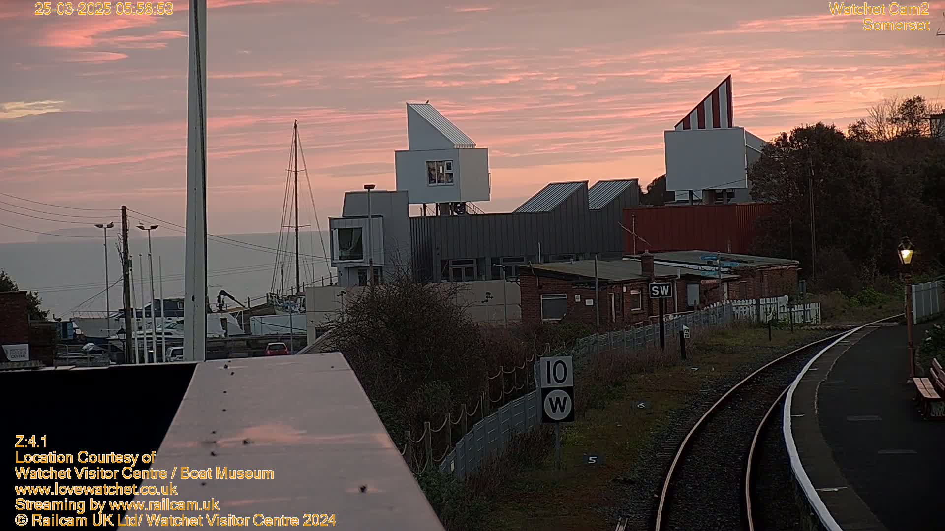 A pink sunrise illuminates a harbor scene with modern buildings, a train track, and boats in the distance.