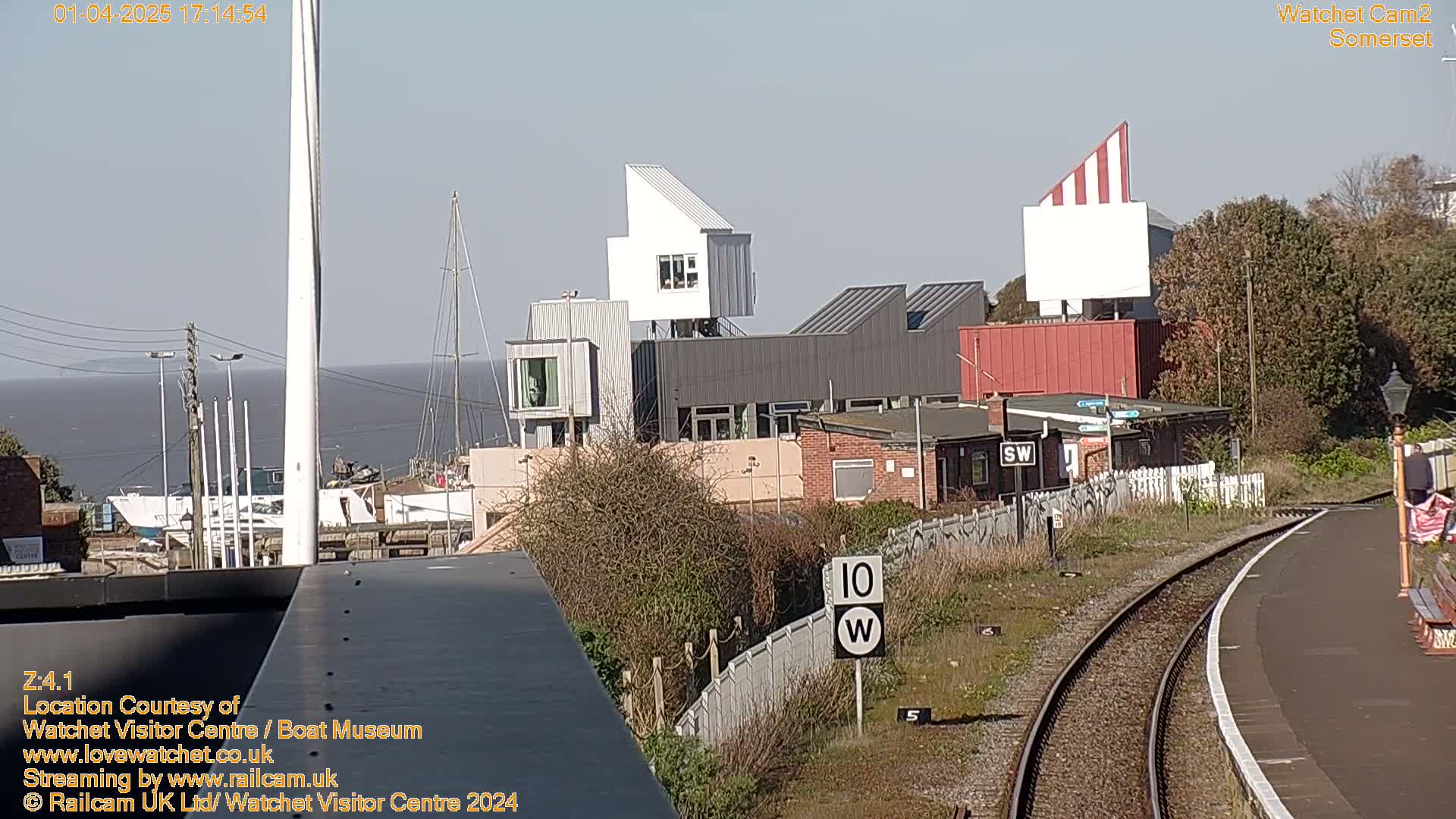 A sunny view of a harbor with modern buildings, boats, and a railway track in the foreground.