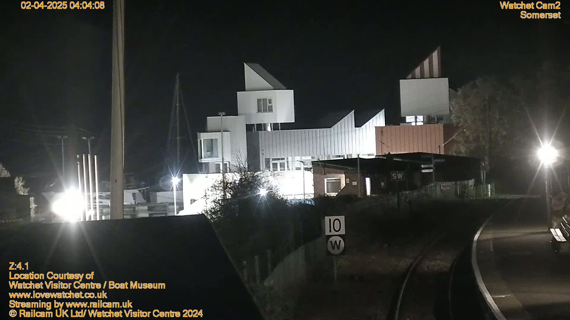 At night, a modern, multi-story building is illuminated, situated near a railway track and a speed limit sign.