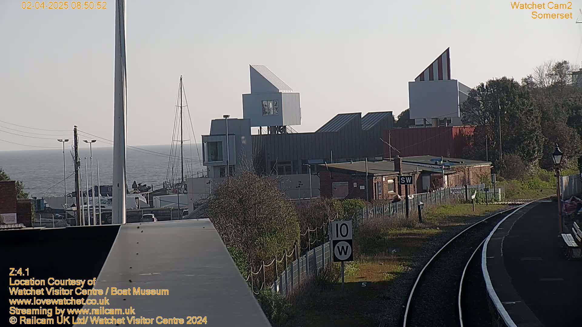A seaside town with modern buildings, a calm ocean, and train tracks is visible under a hazy sky.