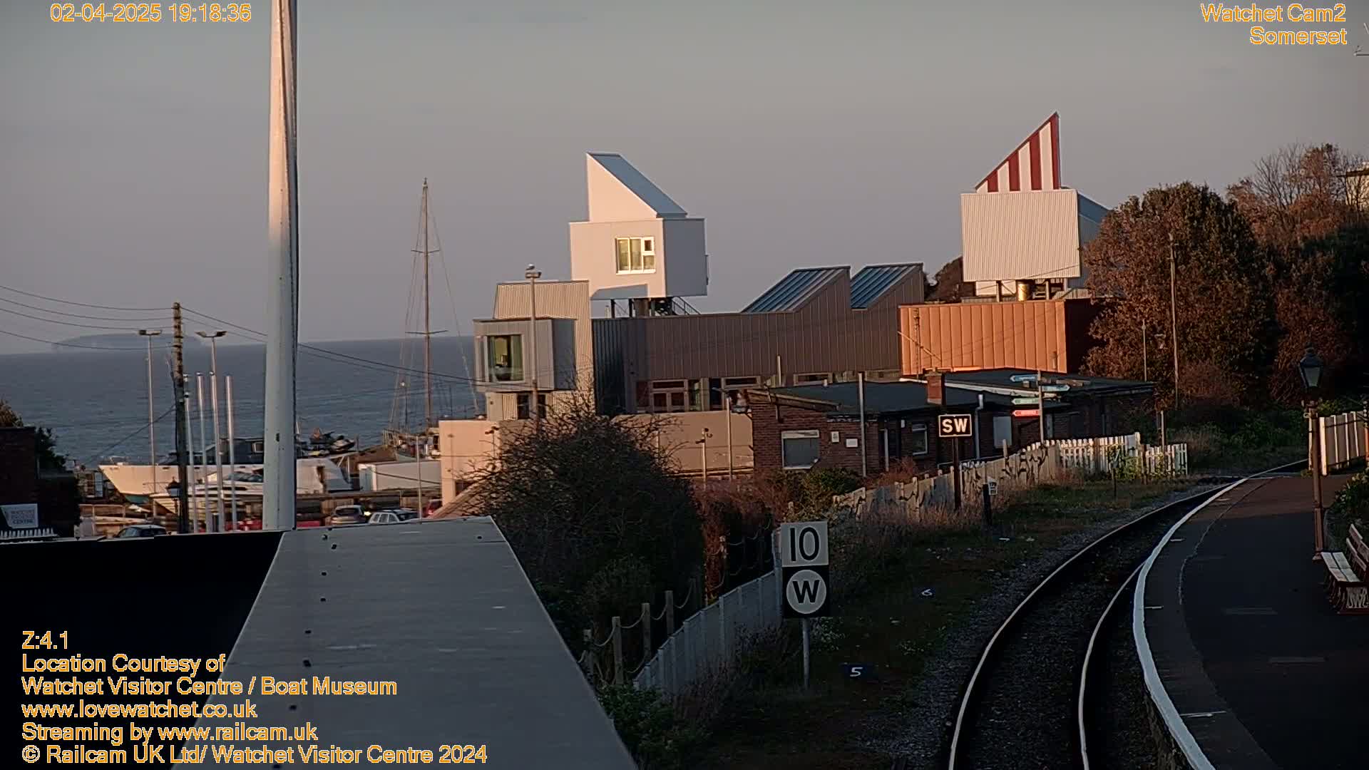 A coastal scene at dusk shows a collection of modern buildings near a train track and harbor with boats, under a clear sky.