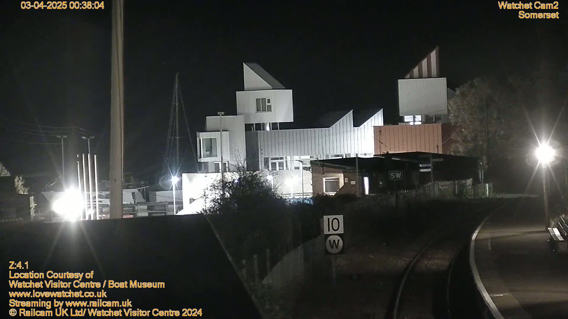 At night, a modern, angular building complex is illuminated, situated next to railroad tracks.