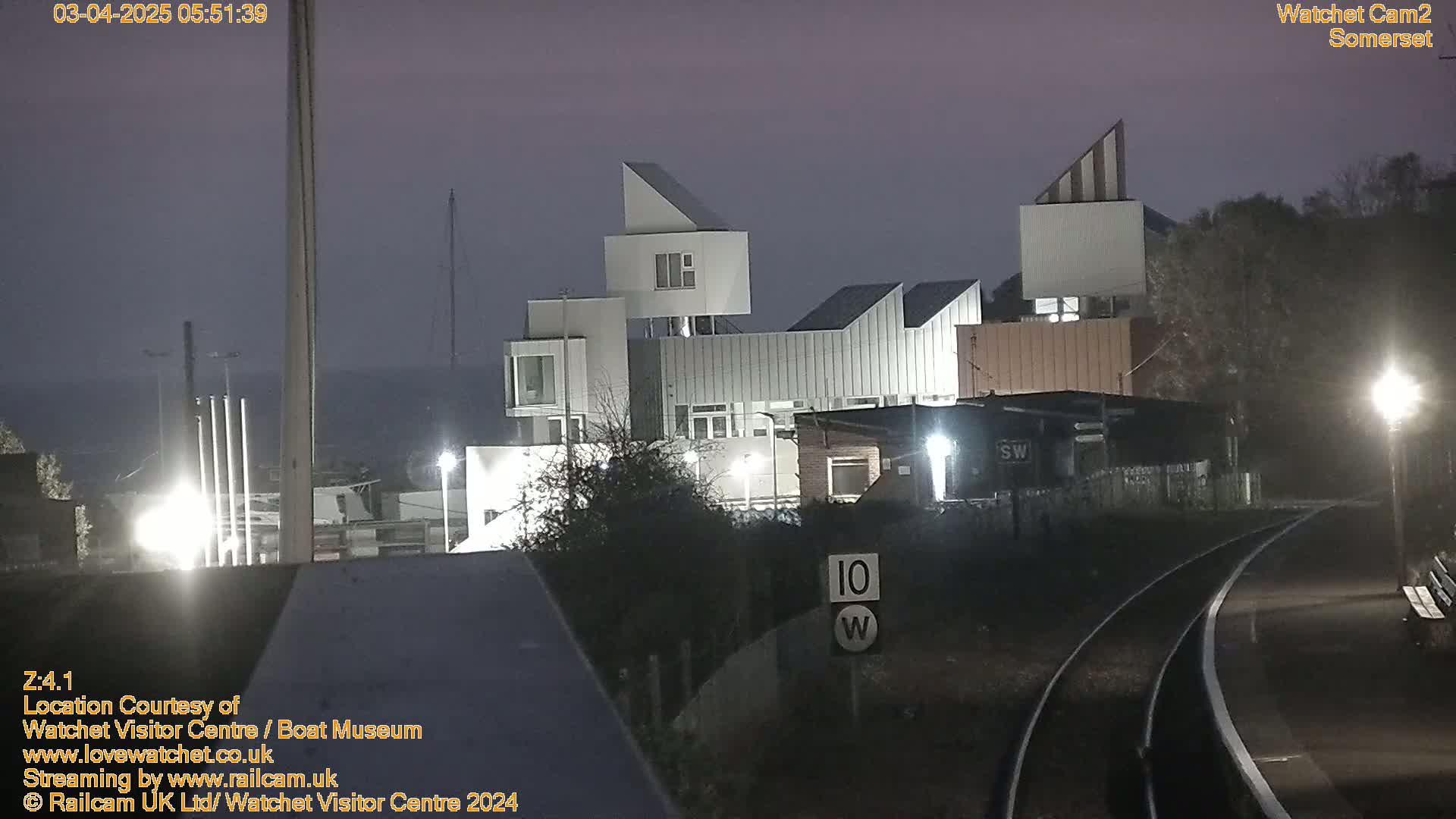 A modern, angular building is illuminated at night under a dusky sky, next to a train track with a speed limit sign.