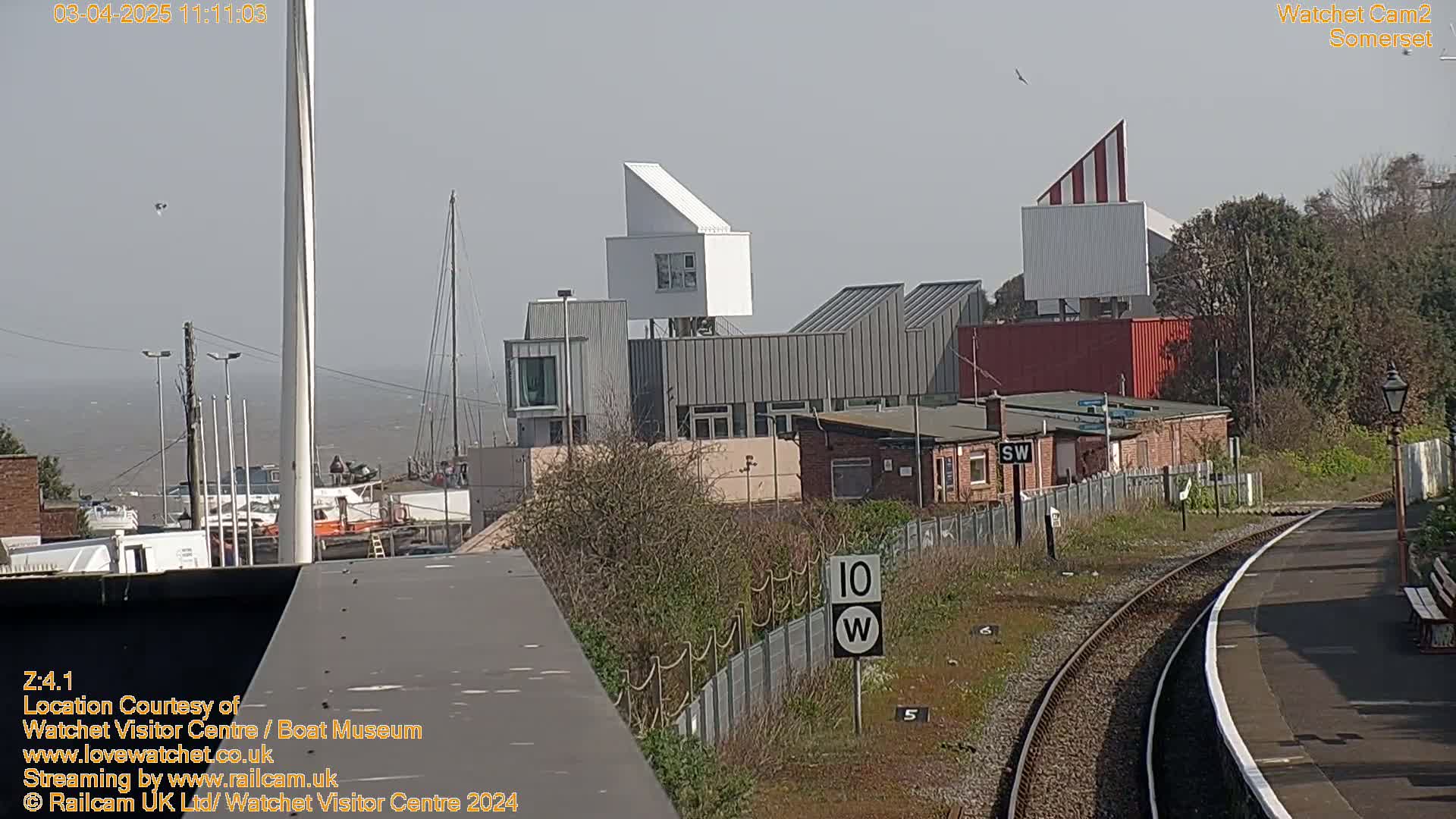 A hazy, sunny day reveals a harbor with boats and buildings, including a modern white structure, near a train station with tracks and a platform.