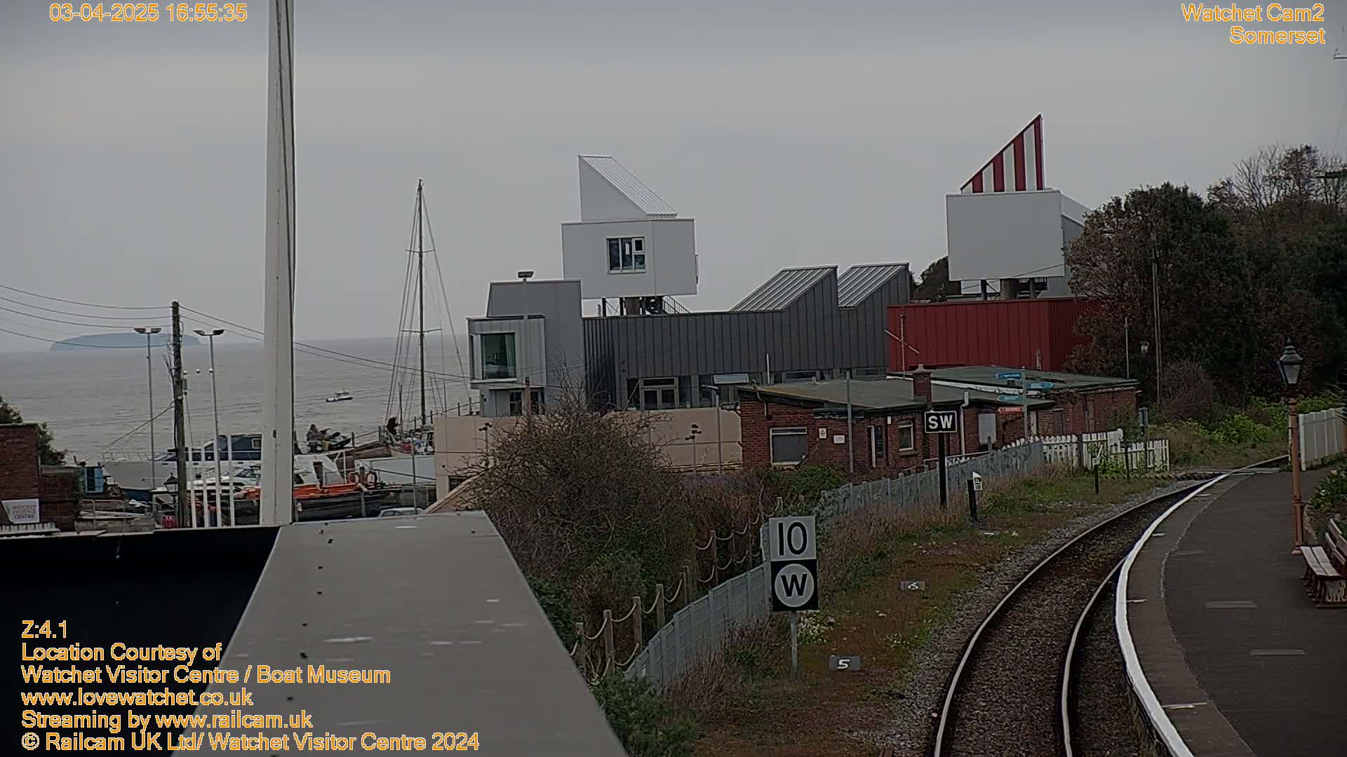 A modern building complex near a train station overlooks a calm body of water under an overcast sky.