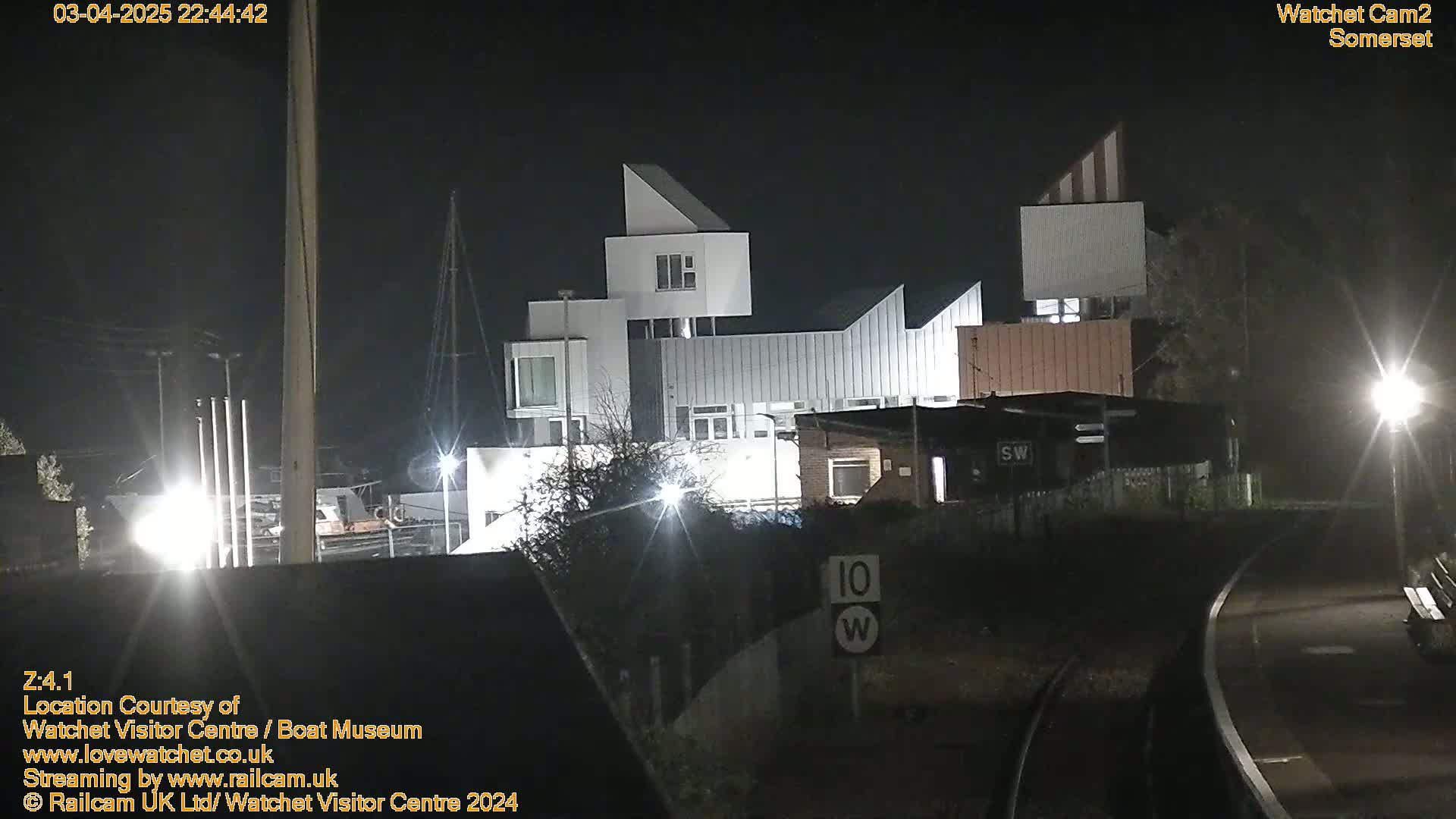 At night, a modern, multi-sectioned building is brightly lit near railroad tracks.