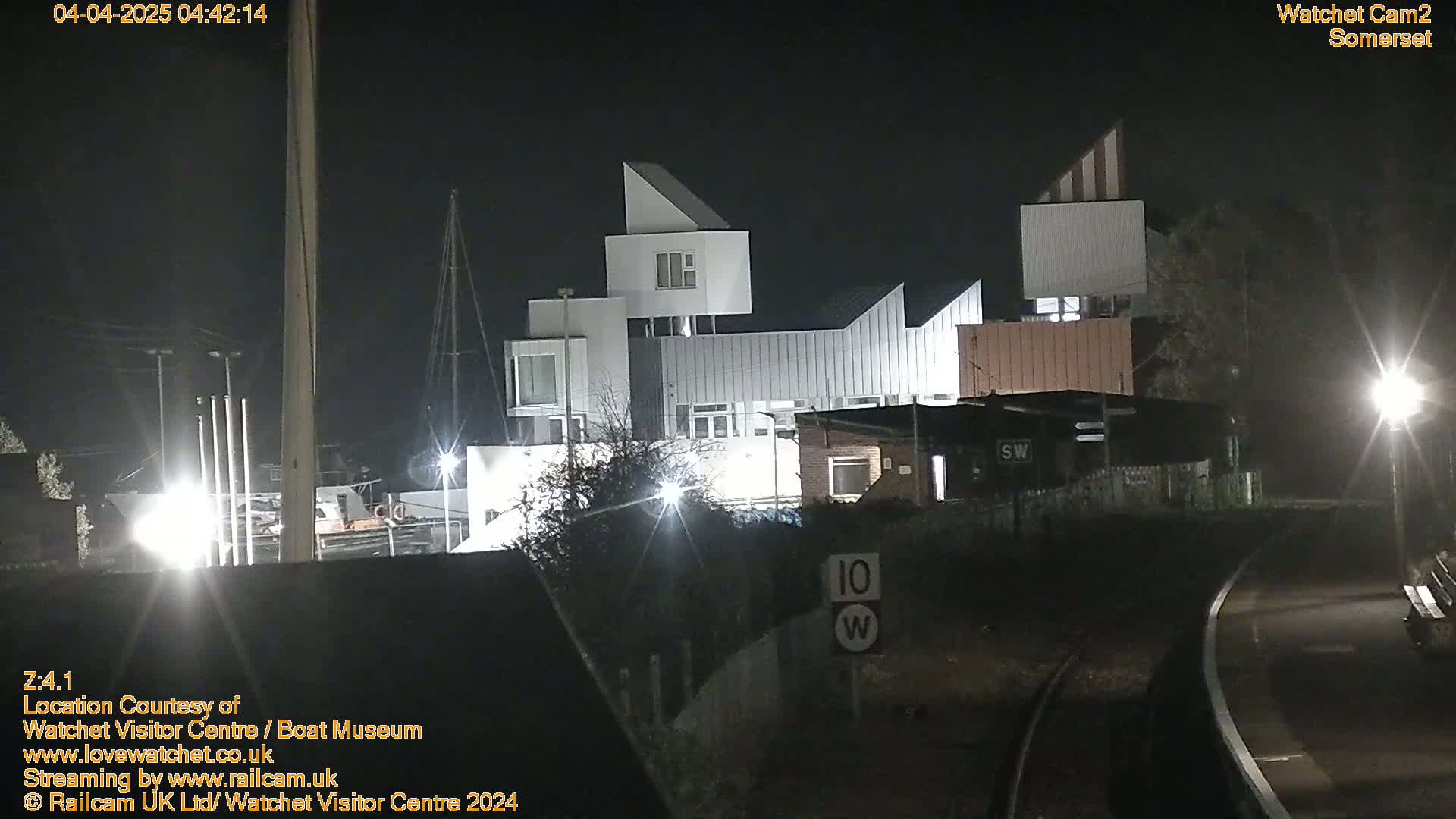 At night, a modern, angular white building complex is illuminated, situated near railroad tracks and some trees.