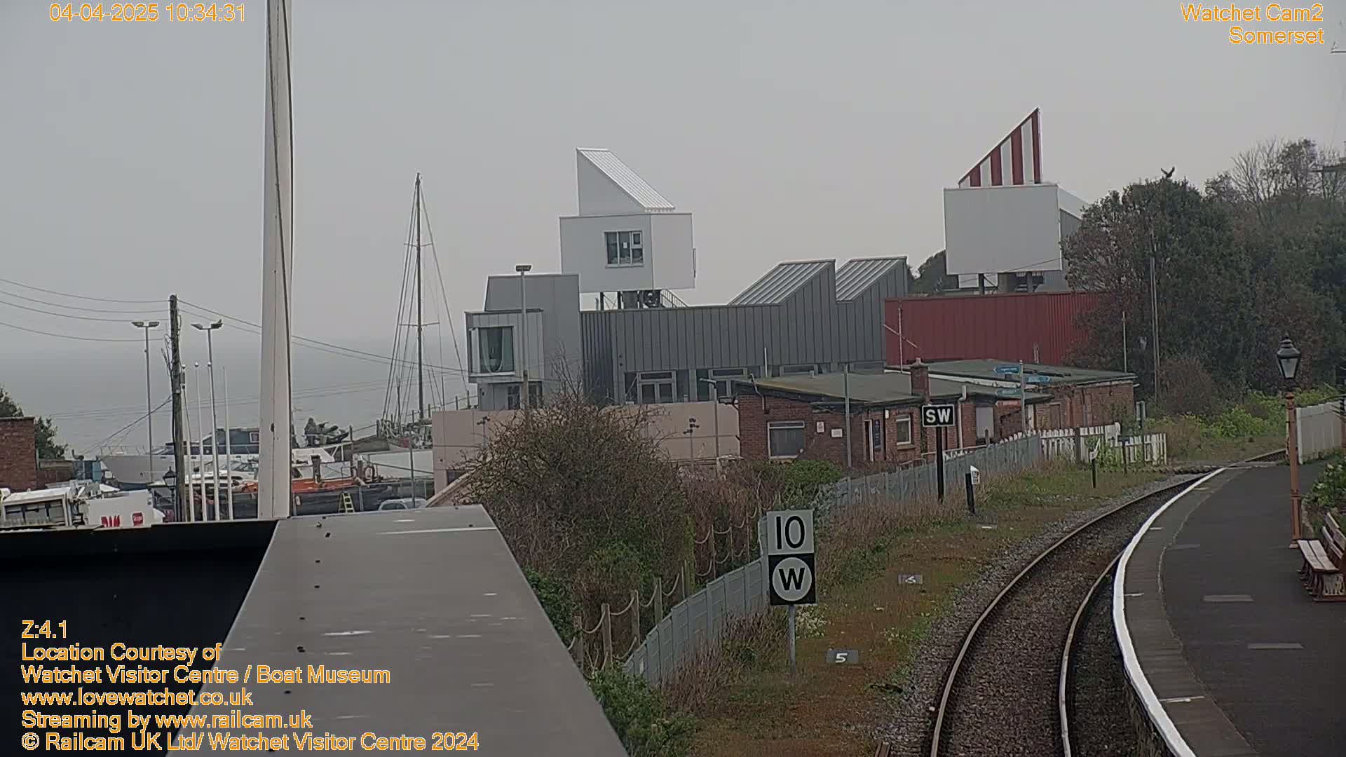 A modern building complex with gray and white siding is seen from across a railway track on an overcast day.