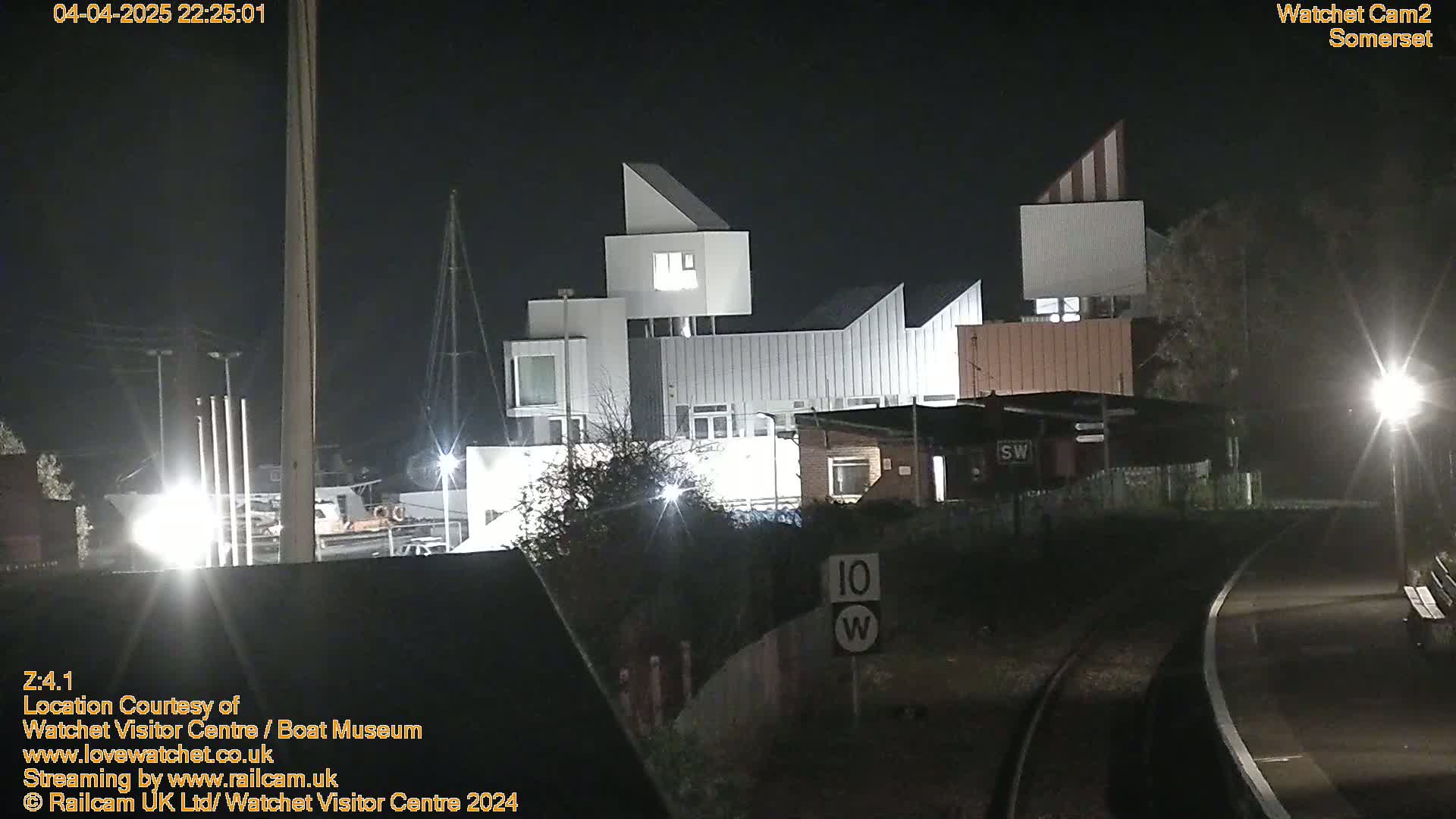 At night, a modern white building is illuminated, situated near a railway track and some low-lying vegetation.