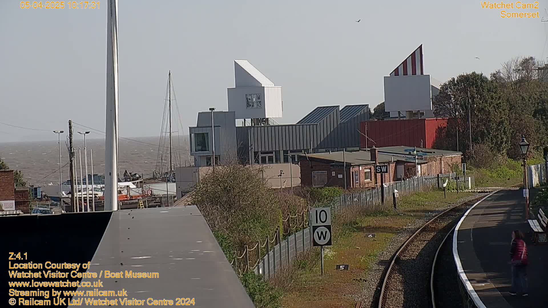 A sunny view of a harbor with modern buildings, boats, and a train track, with a person standing on the platform.