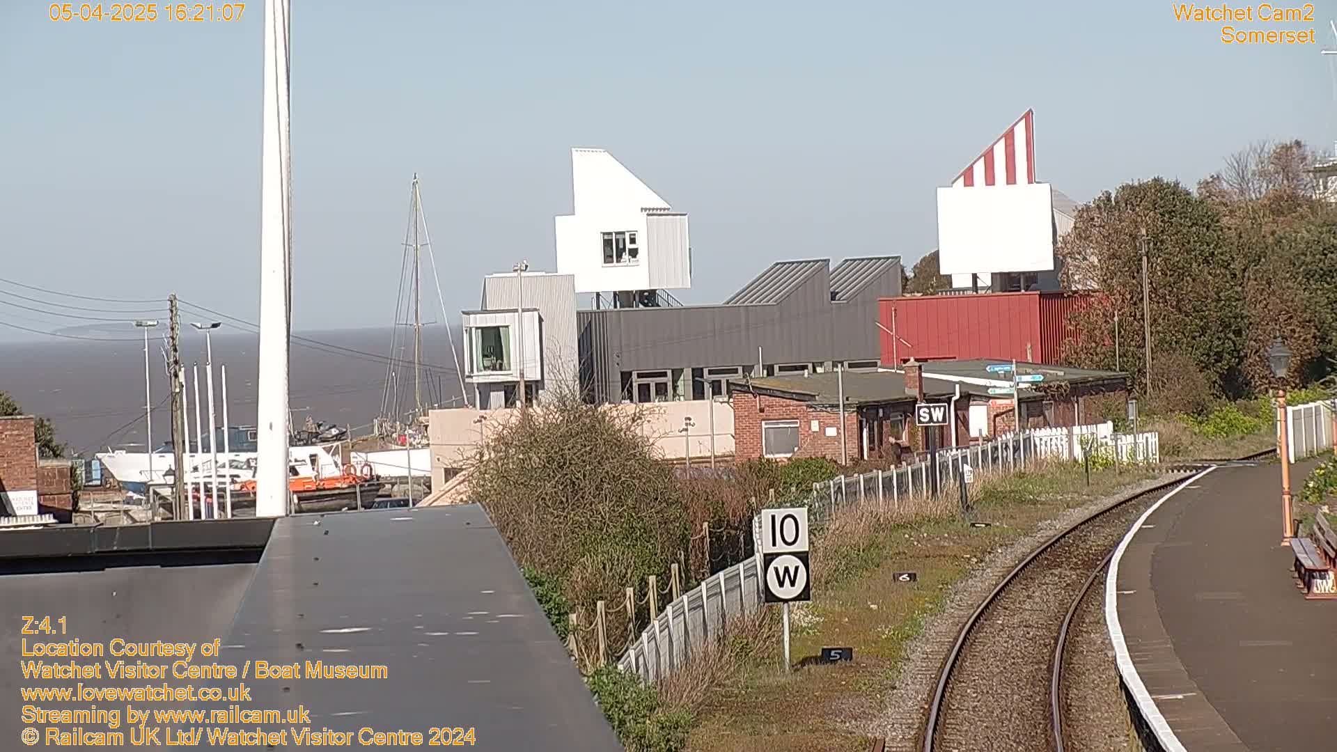 A waterfront scene on a sunny day shows a modern building complex near a harbor with boats, and railway tracks curving into the distance.
