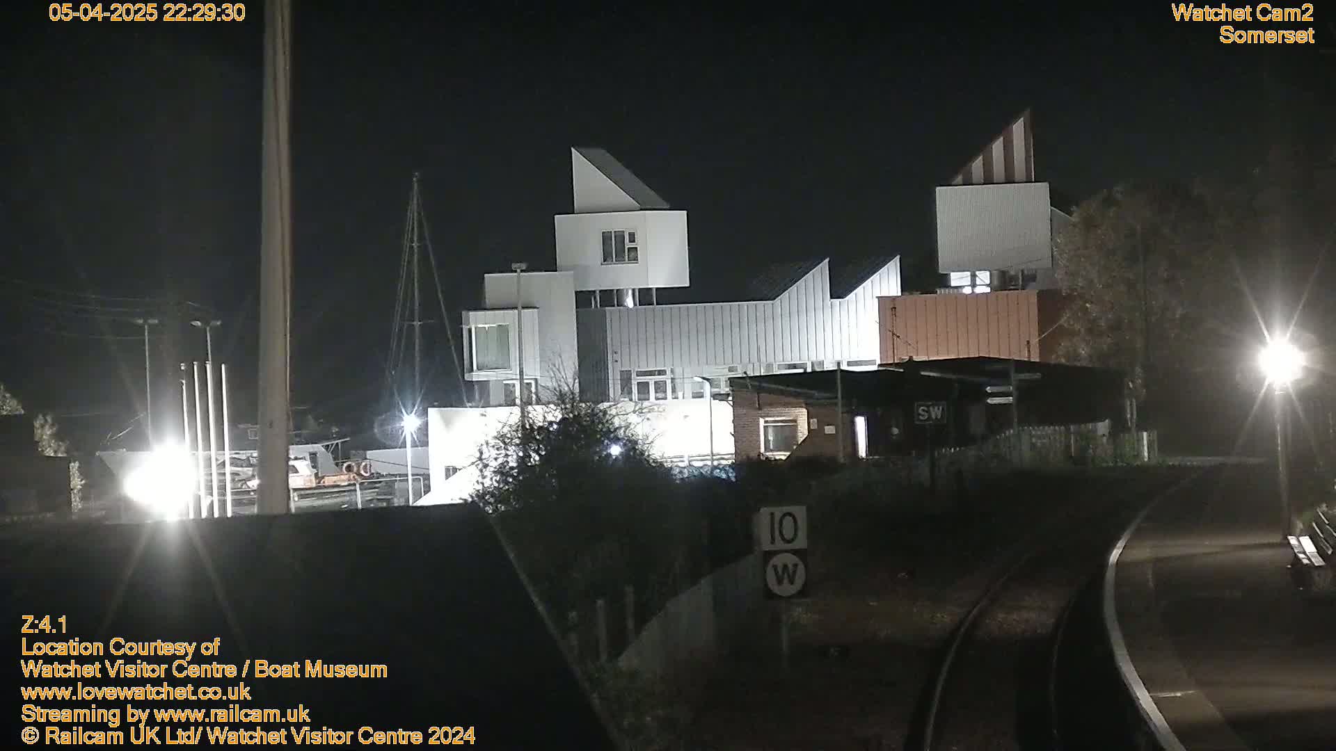 At night, a modern, multi-sectioned building is illuminated, situated near a railway track with a speed limit sign.