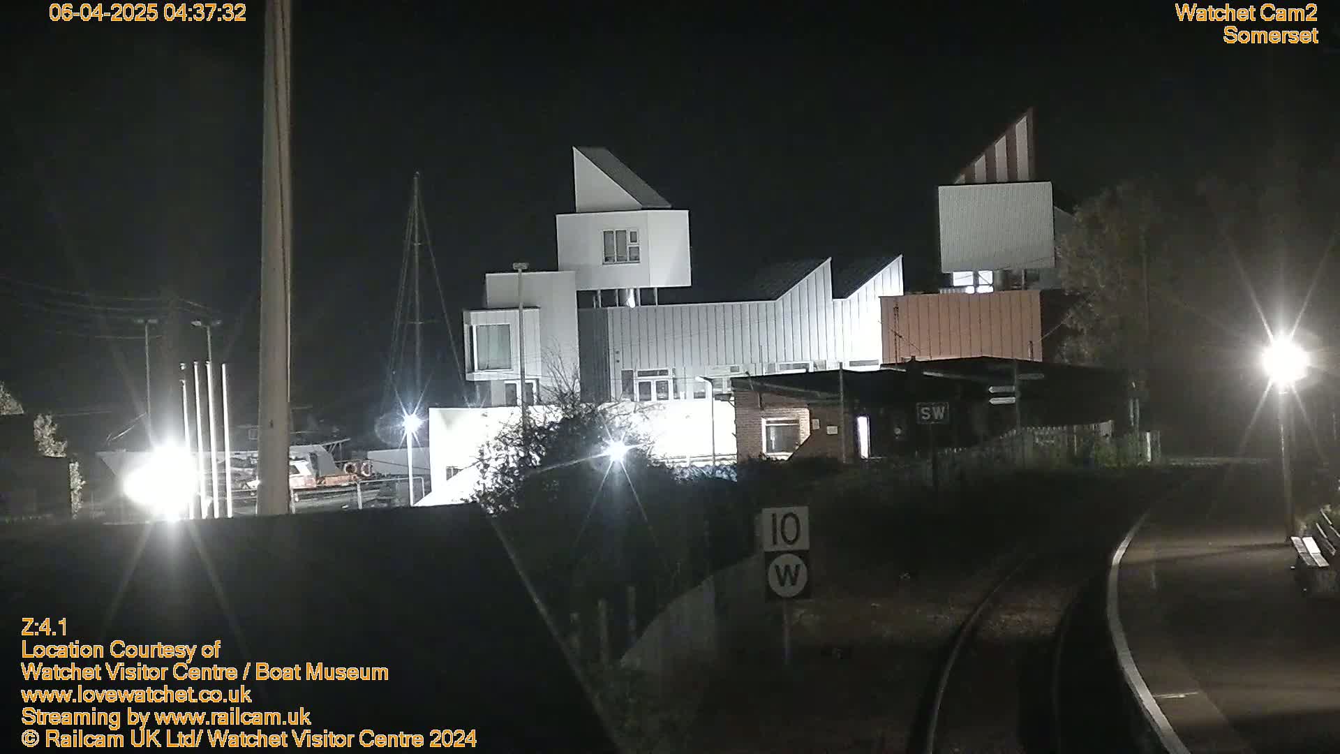 At night, a modern, multi-story building is illuminated, adjacent to railroad tracks and a streetlight.