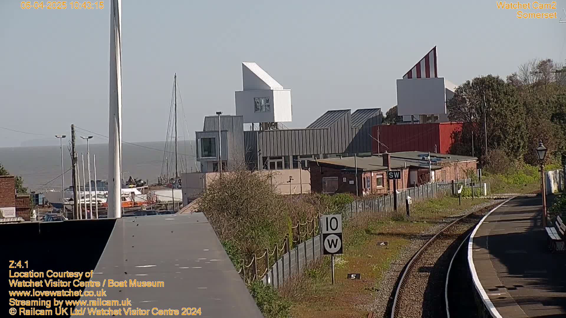 A modern building complex near a train station and harbor, with boats visible in the distance, on a sunny day.