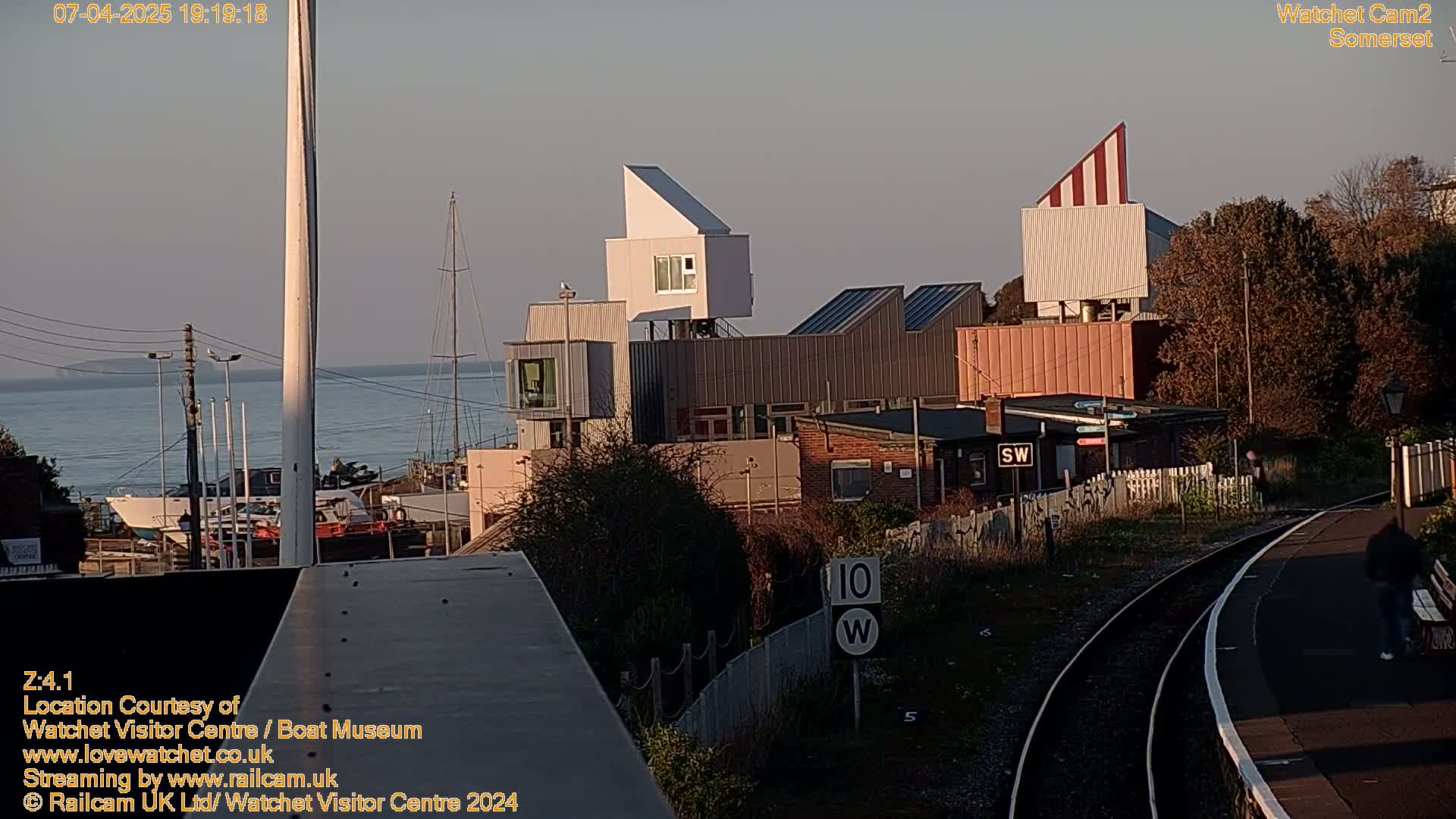 A seaside town at dusk is shown, featuring modern buildings near a harbor with boats and a railway track in the foreground.