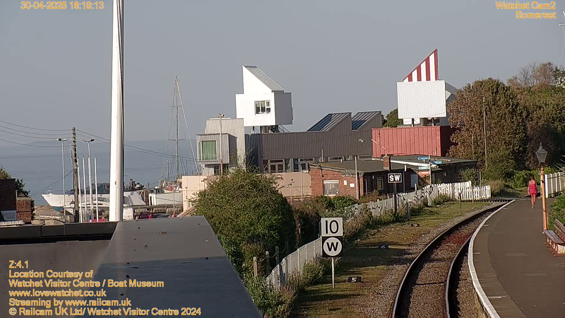 A view from a train shows a harbor with boats, modern buildings, a railway track, and a person walking on a platform under a mostly clear sky.