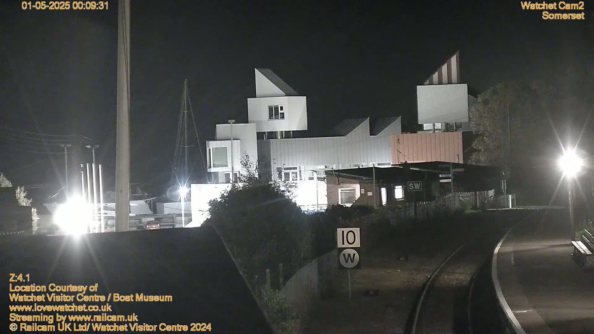 A modern, multi-level building is illuminated at night near a railway track, under a clear, dark sky.