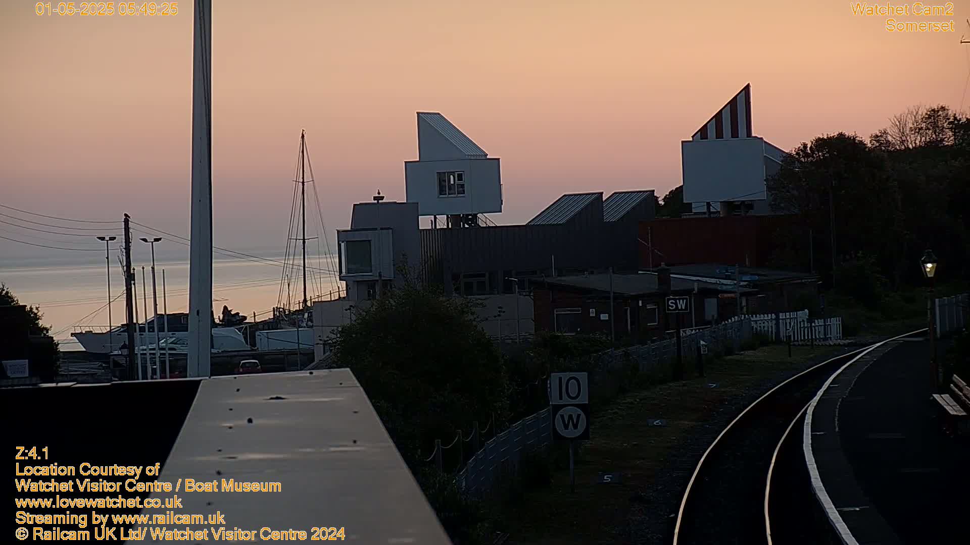 A pink sunrise illuminates a harbor with modern buildings and a railway line in the foreground.