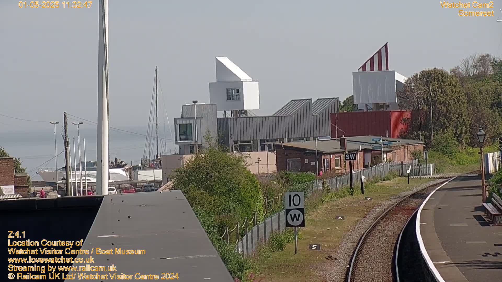 A modern building complex overlooks a harbor with boats, under a bright, sunny sky, near a railway track.