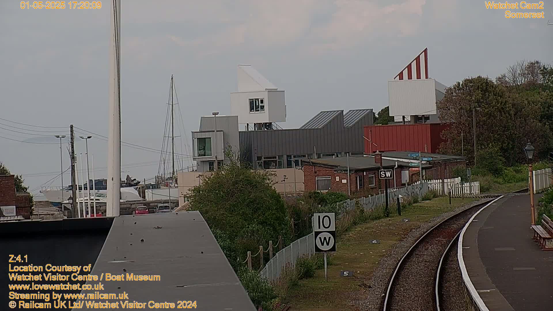 A view of a modern building complex near a train station on an overcast day, with boats visible in the background.