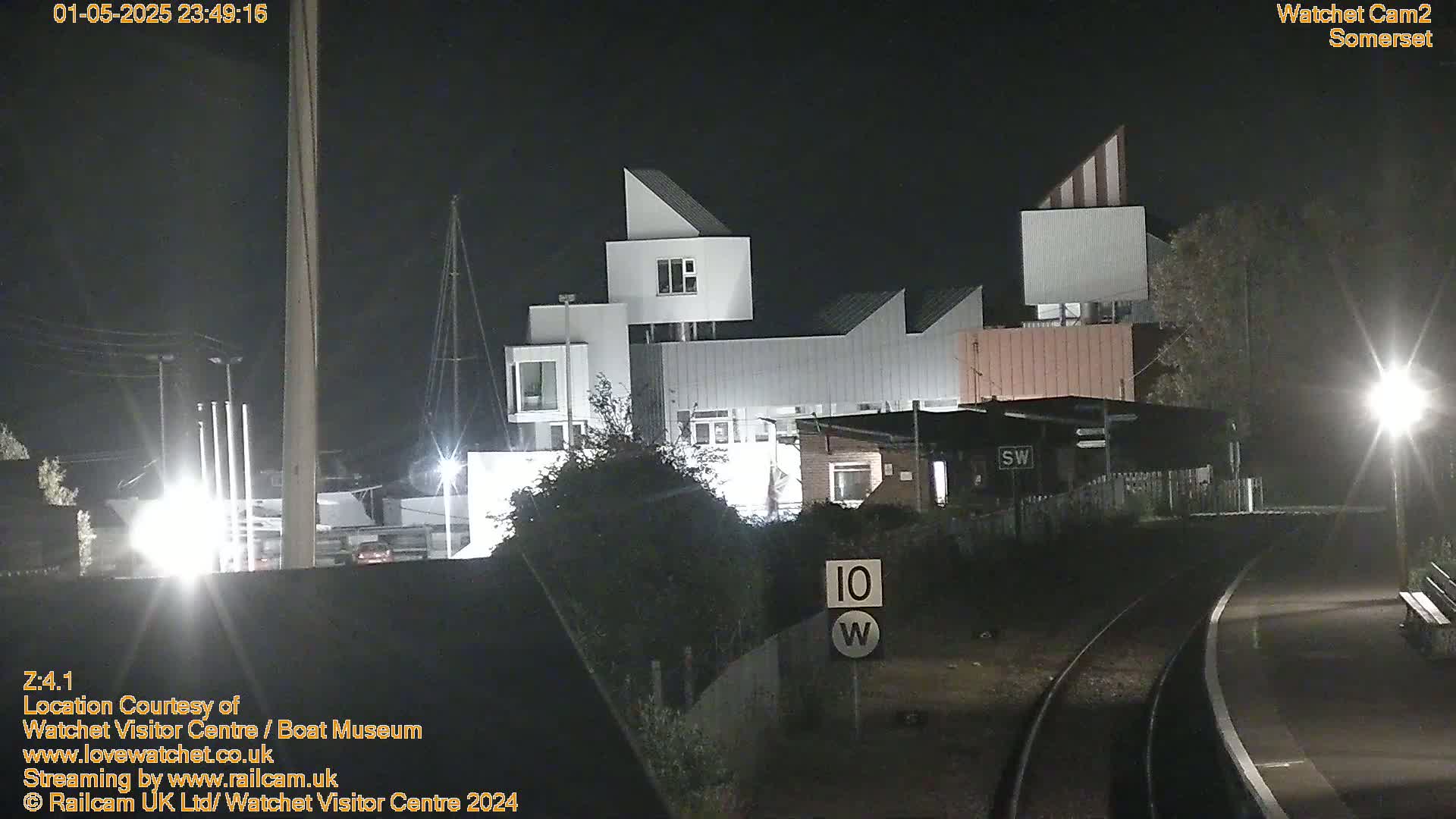 A modern, angular building complex is illuminated at night near train tracks, under a dark sky.