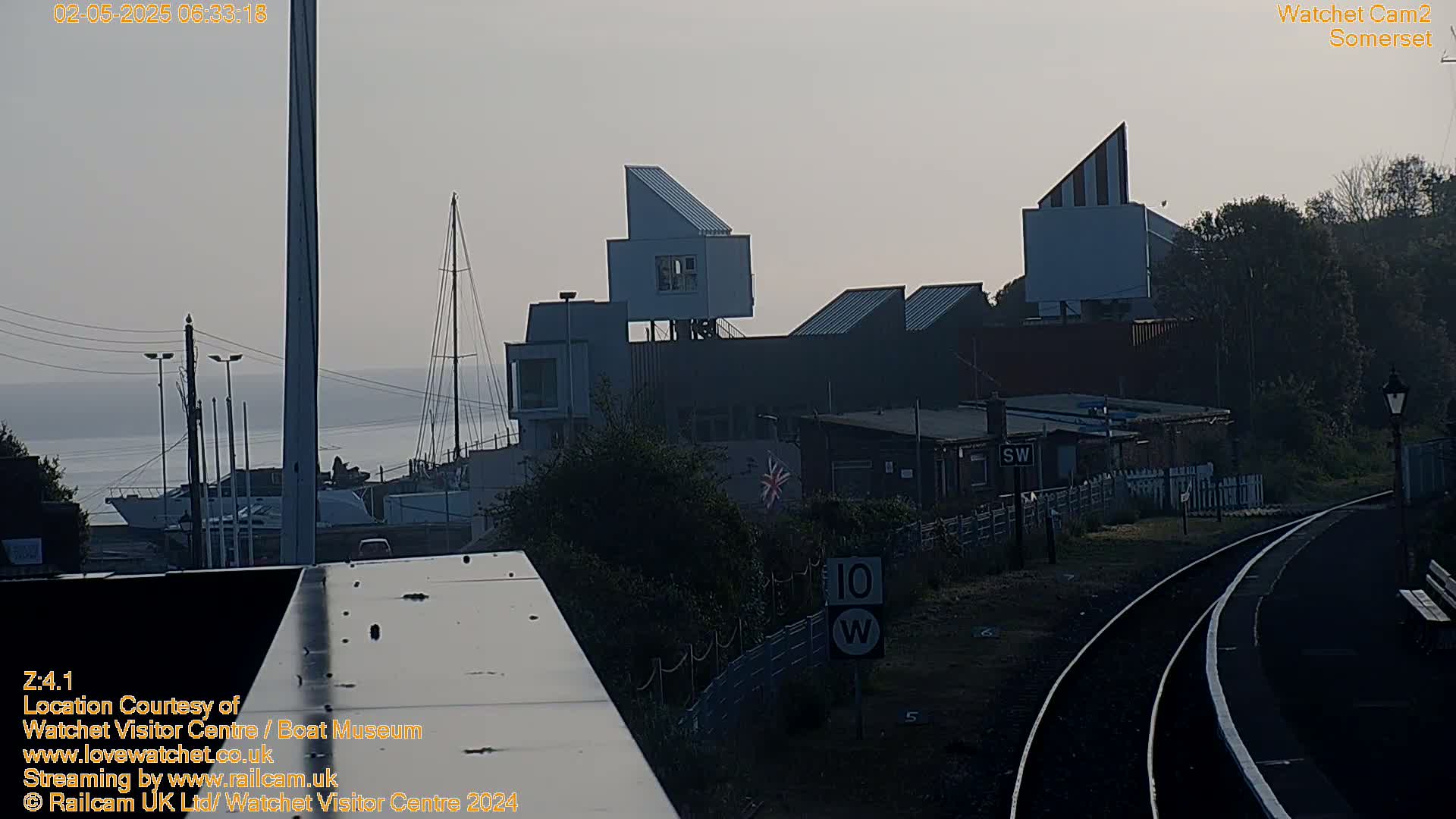 A hazy sunrise illuminates a harbor with boats, a modern building complex, and railway tracks curving into the distance.