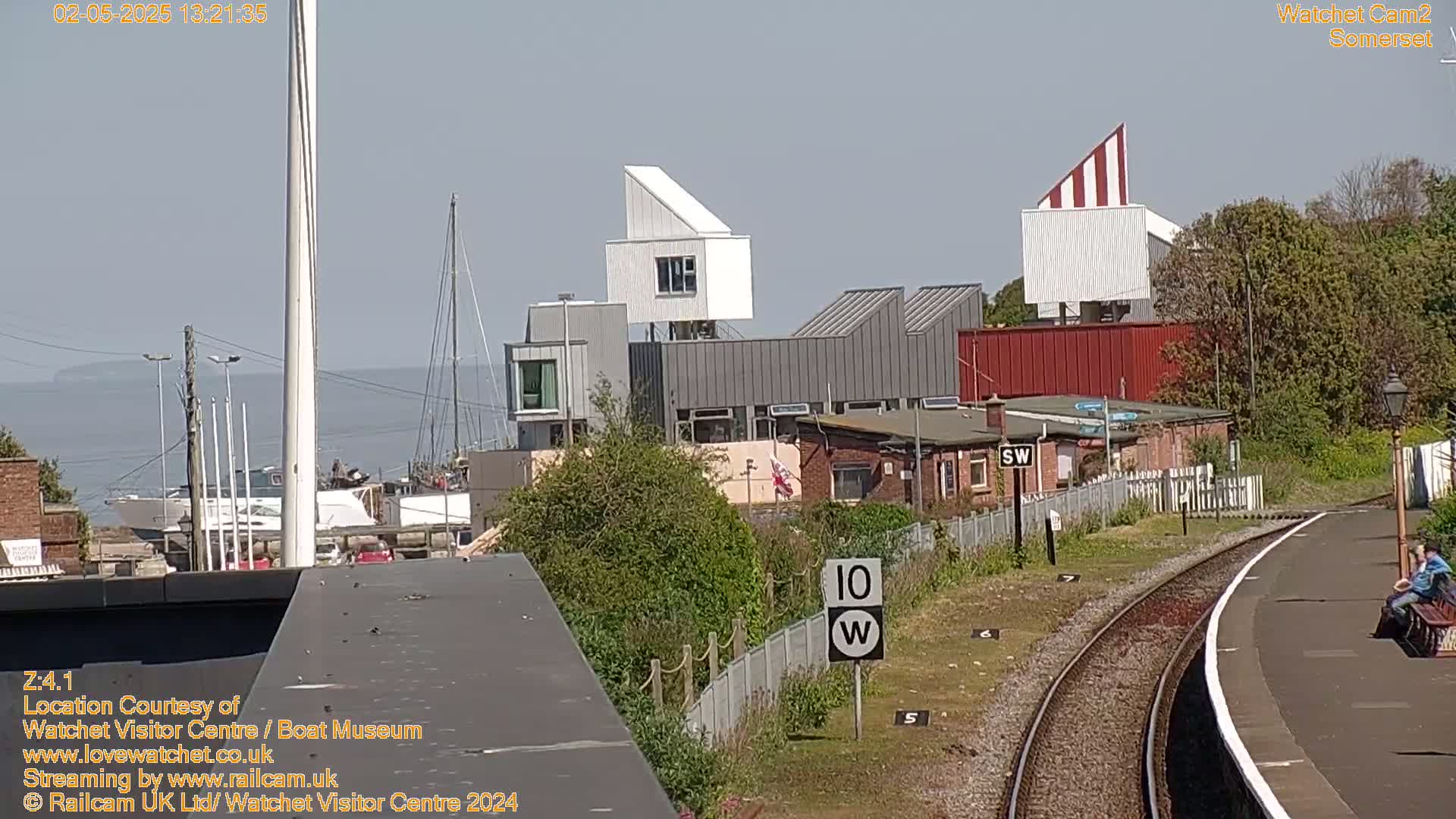 A sunny day overlooks a harbor with boats and modern buildings, including a distinctive white and red striped structure, beside a train station platform with a person sitting on a bench.