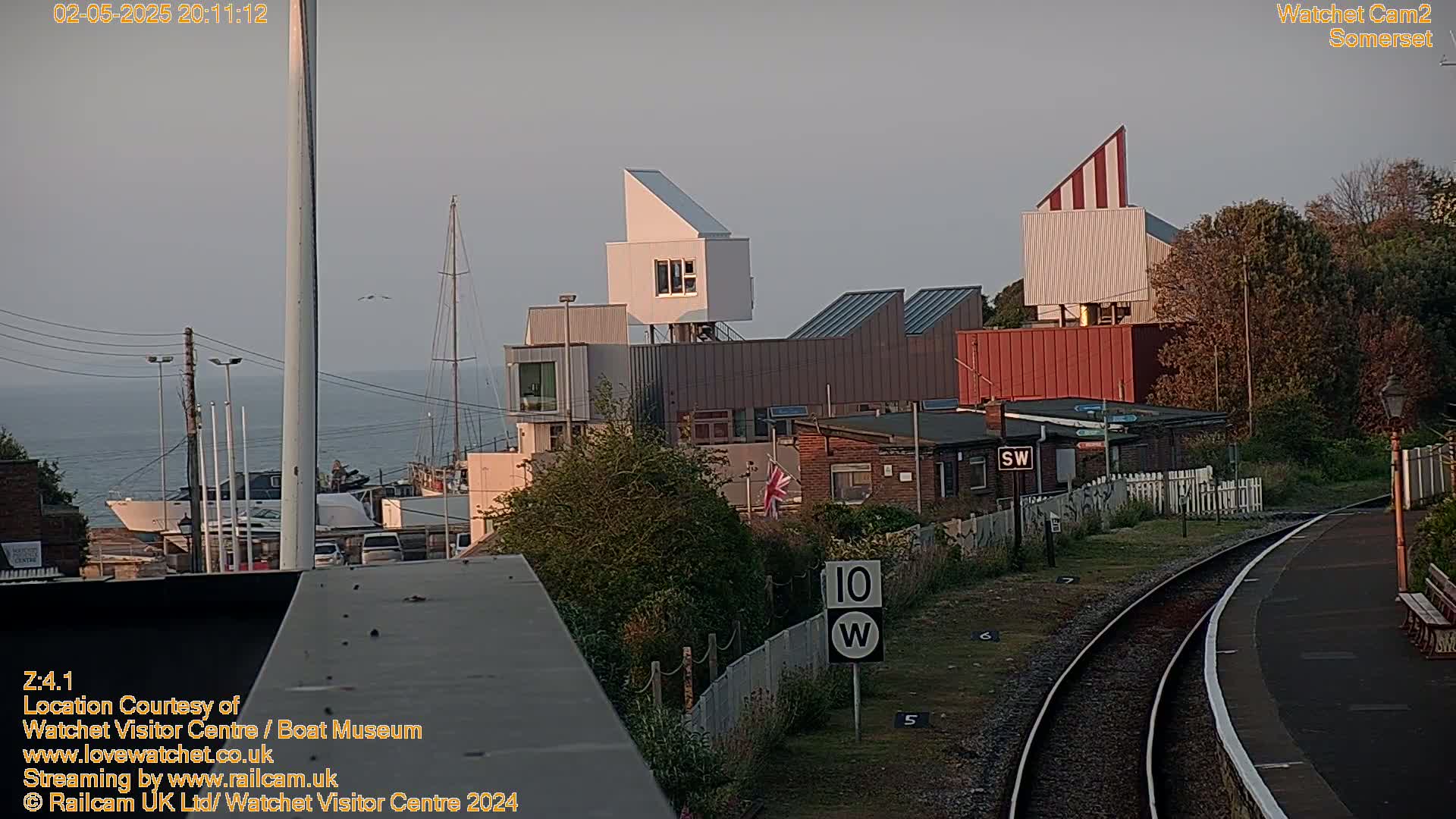 A seaside town at dusk, featuring modern buildings near a harbor, a railway track, and calm water.