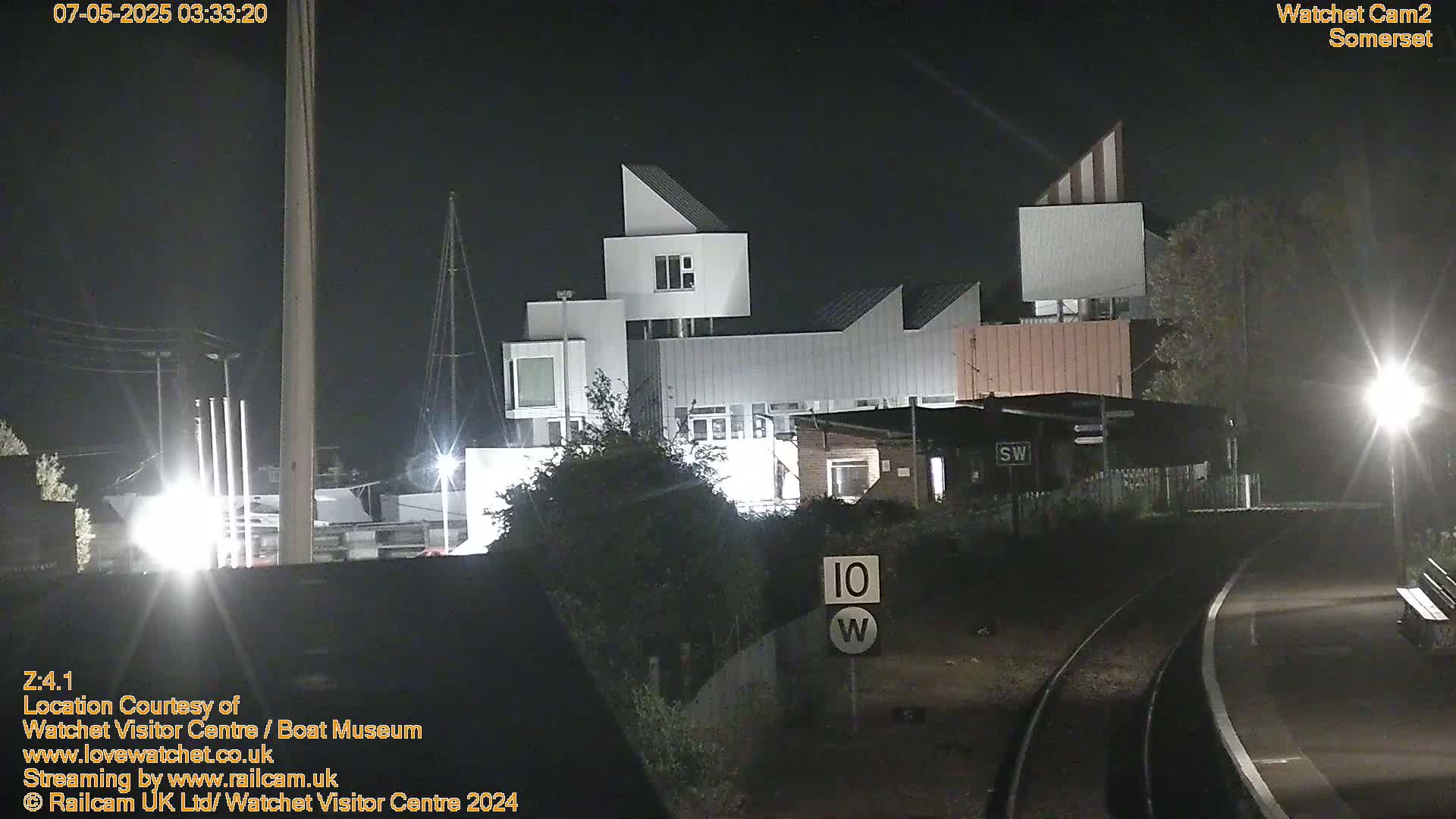 A modern, multi-level white building is illuminated at night near railroad tracks.