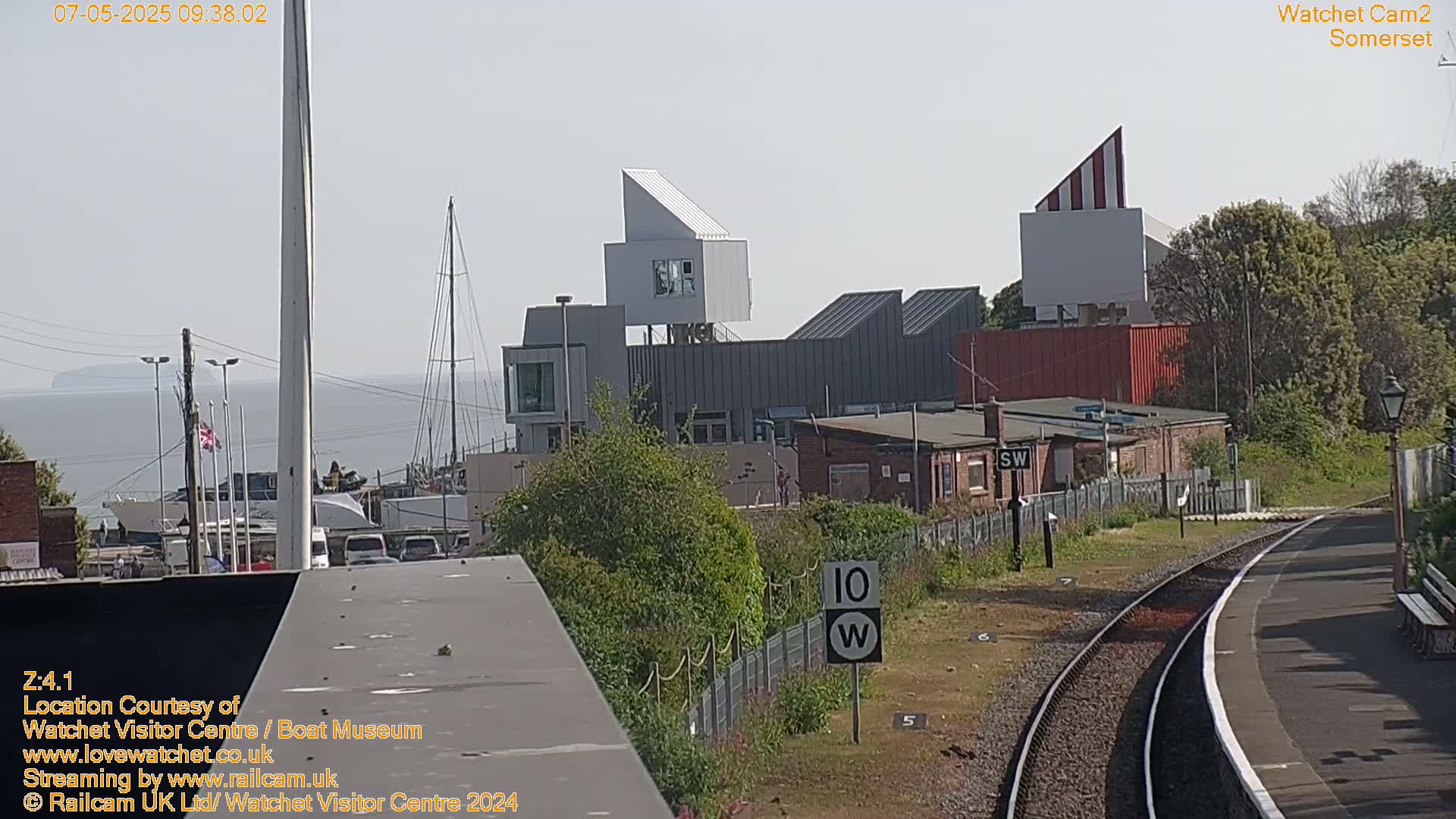 A waterfront view on a sunny day shows modern buildings, a train track, and a calm body of water with a distant island visible in the background.