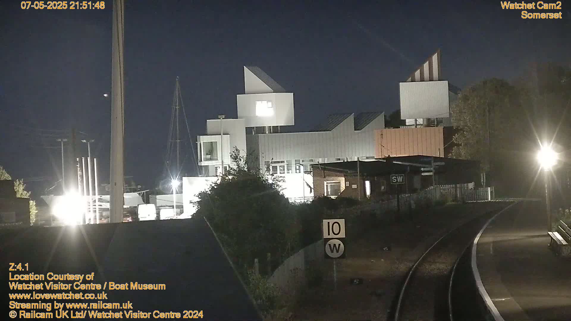 At night, a modern building complex is illuminated, with train tracks and a railway platform visible in the foreground.