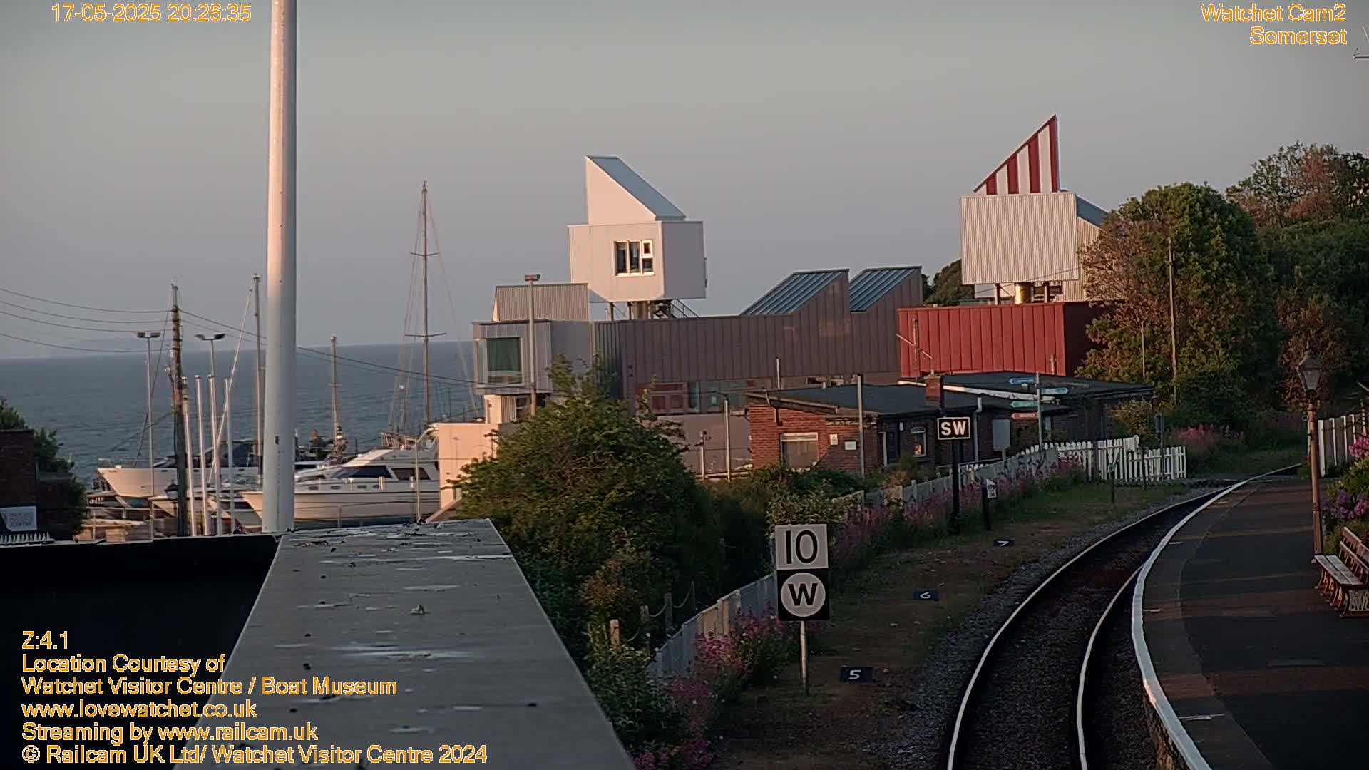 A harbor with boats, modern buildings, and a train track is seen under a mostly clear evening sky.