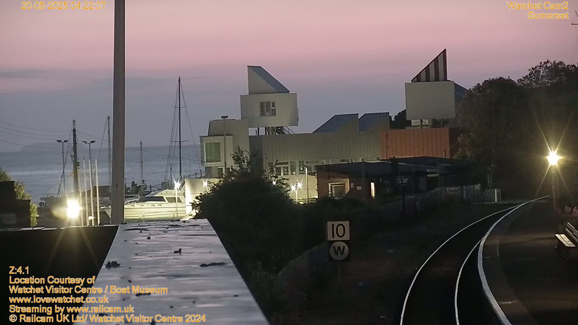 A twilight view of a harbor with boats and modern buildings near a railway track under a pinkish sky.