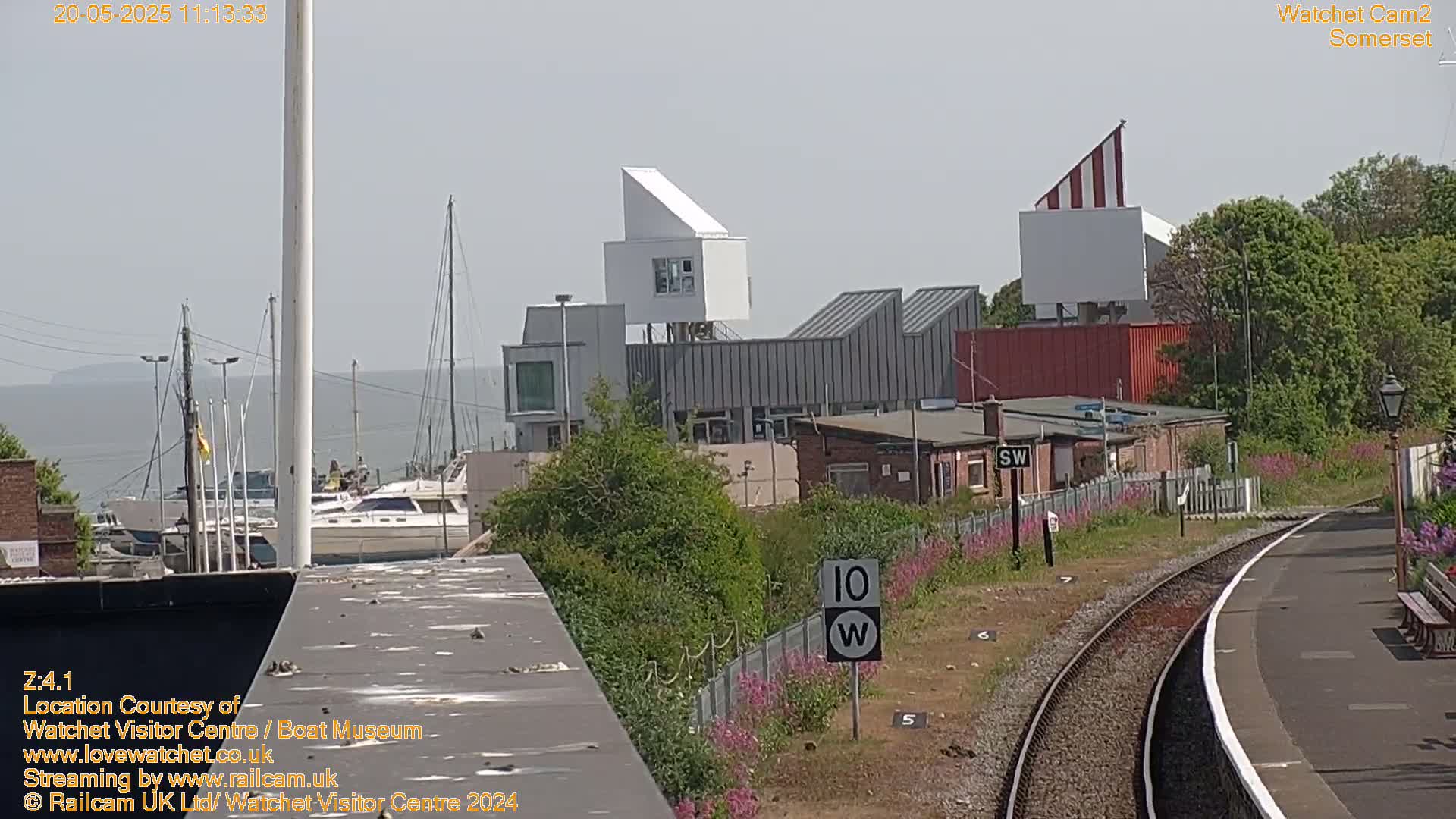 A harbor with boats, modern buildings, and a train track with a speed limit sign is seen on a partly sunny day.