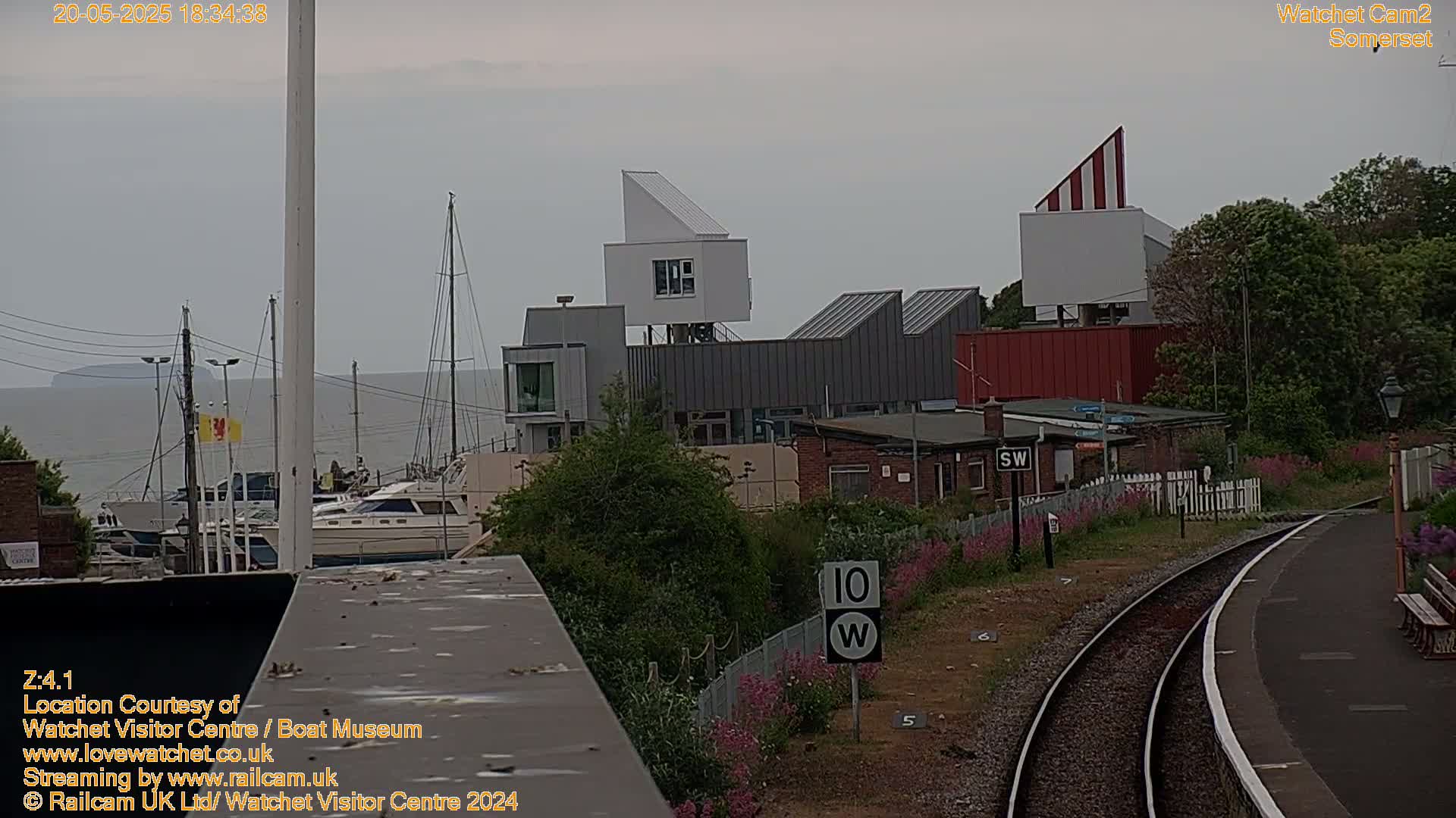 A harbor with boats, modern buildings, and a railway track is seen on an overcast day.