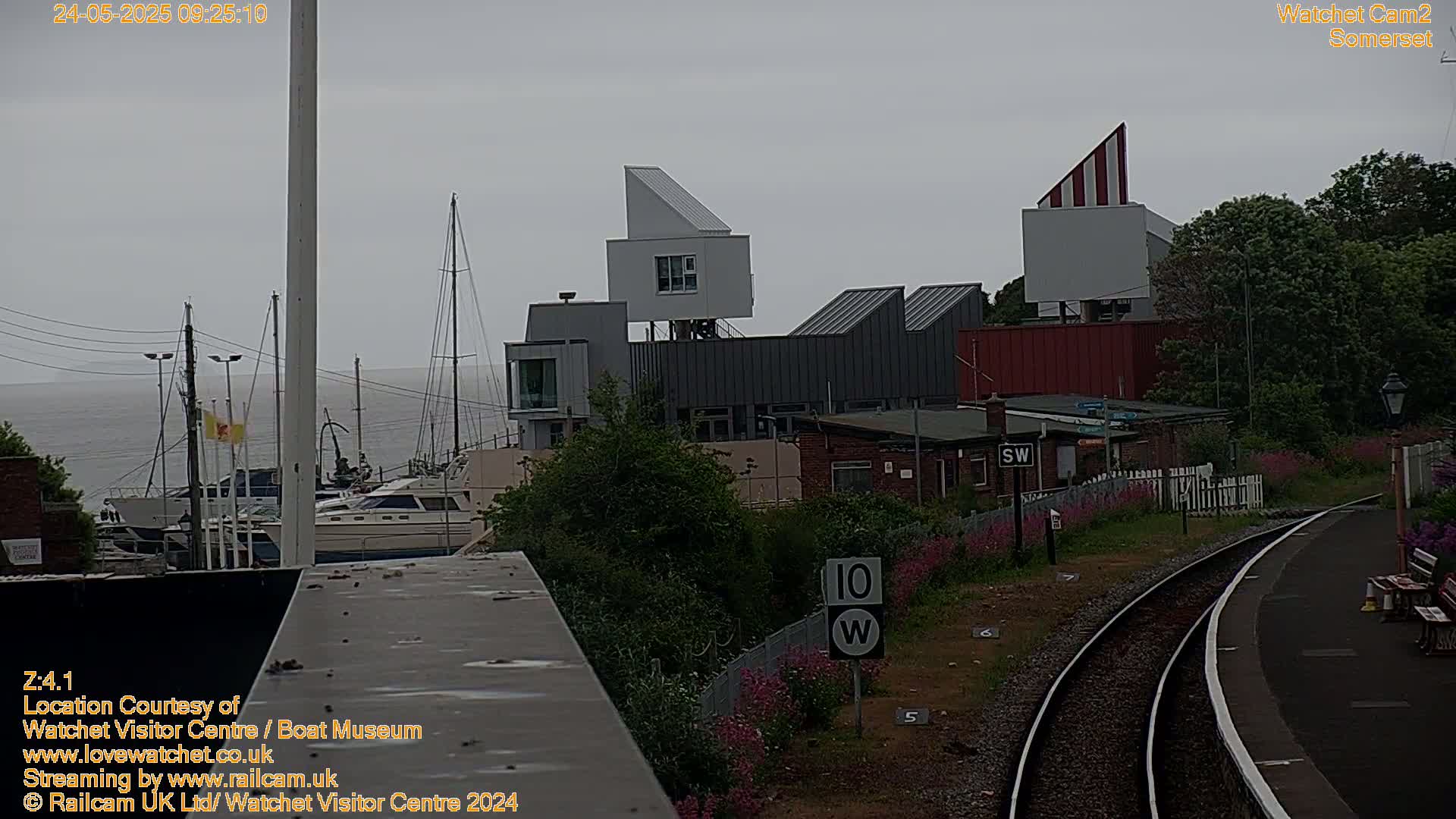 A gray, overcast day shows a marina with boats, modern buildings, and a railway line with pink flowers growing alongside it.