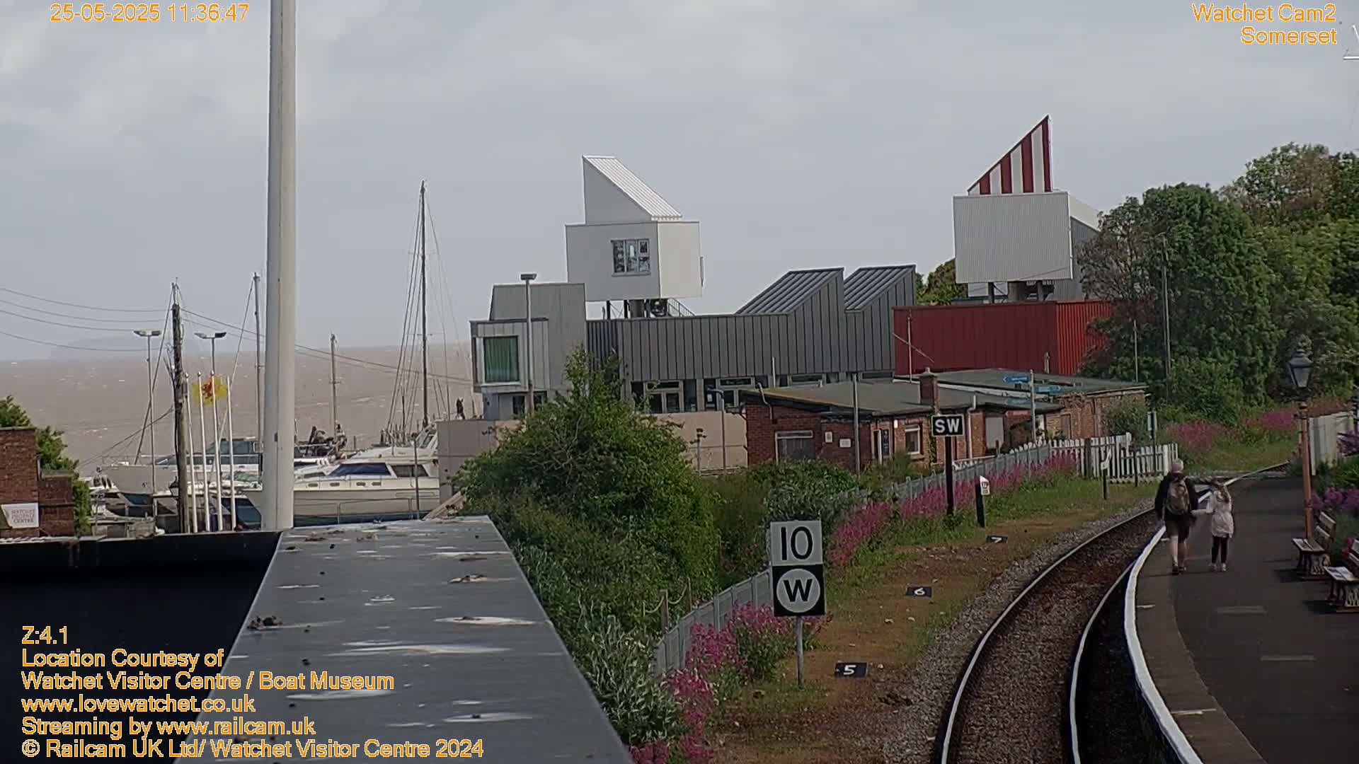 A harbor with docked boats is visible beyond modern buildings and a railway platform where two people are walking.